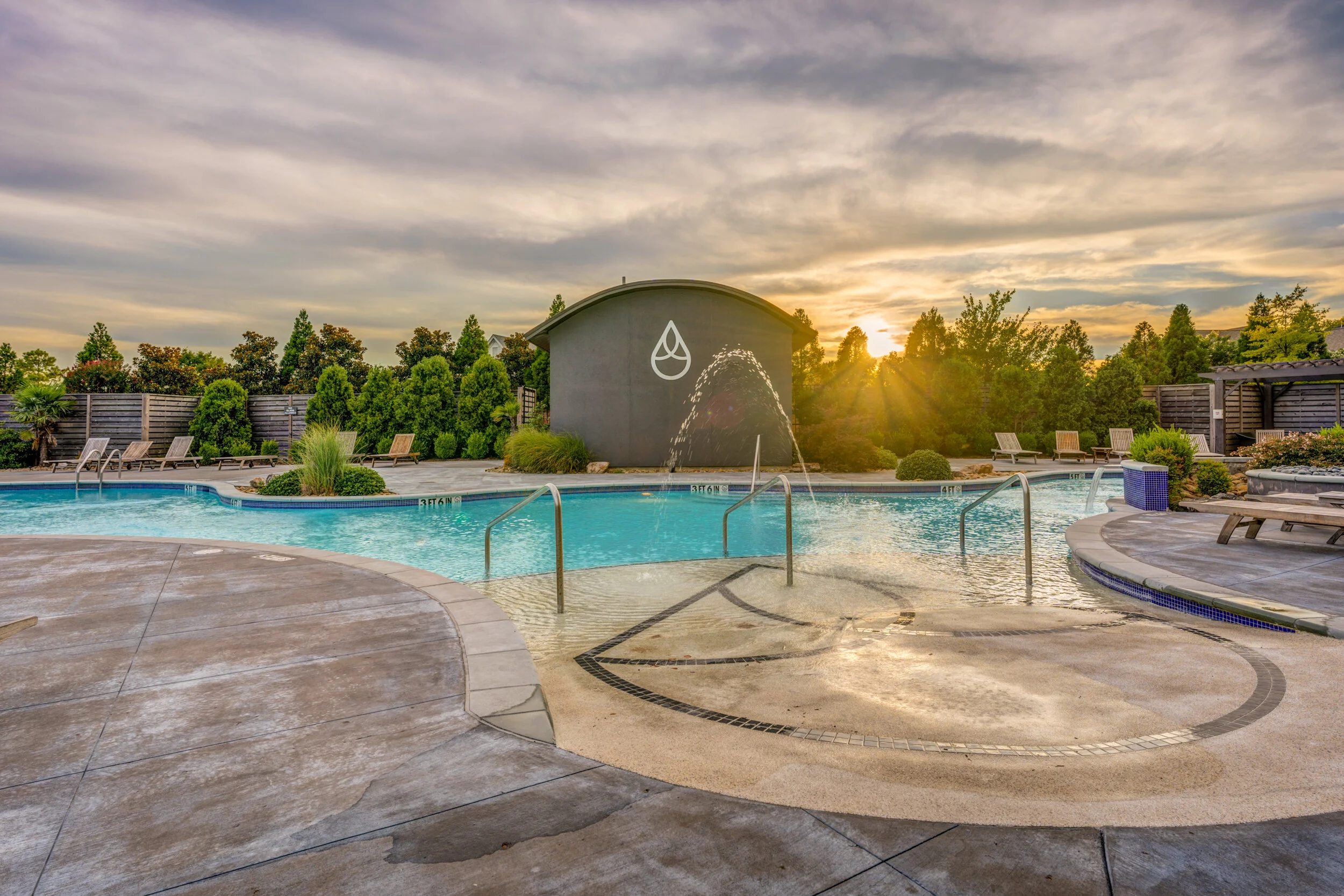 Outdoor swimming pool at sunset with lounge chairs, greenery, and a dark building with a water droplet logo and fountain feature. Memphis real estate photography. 