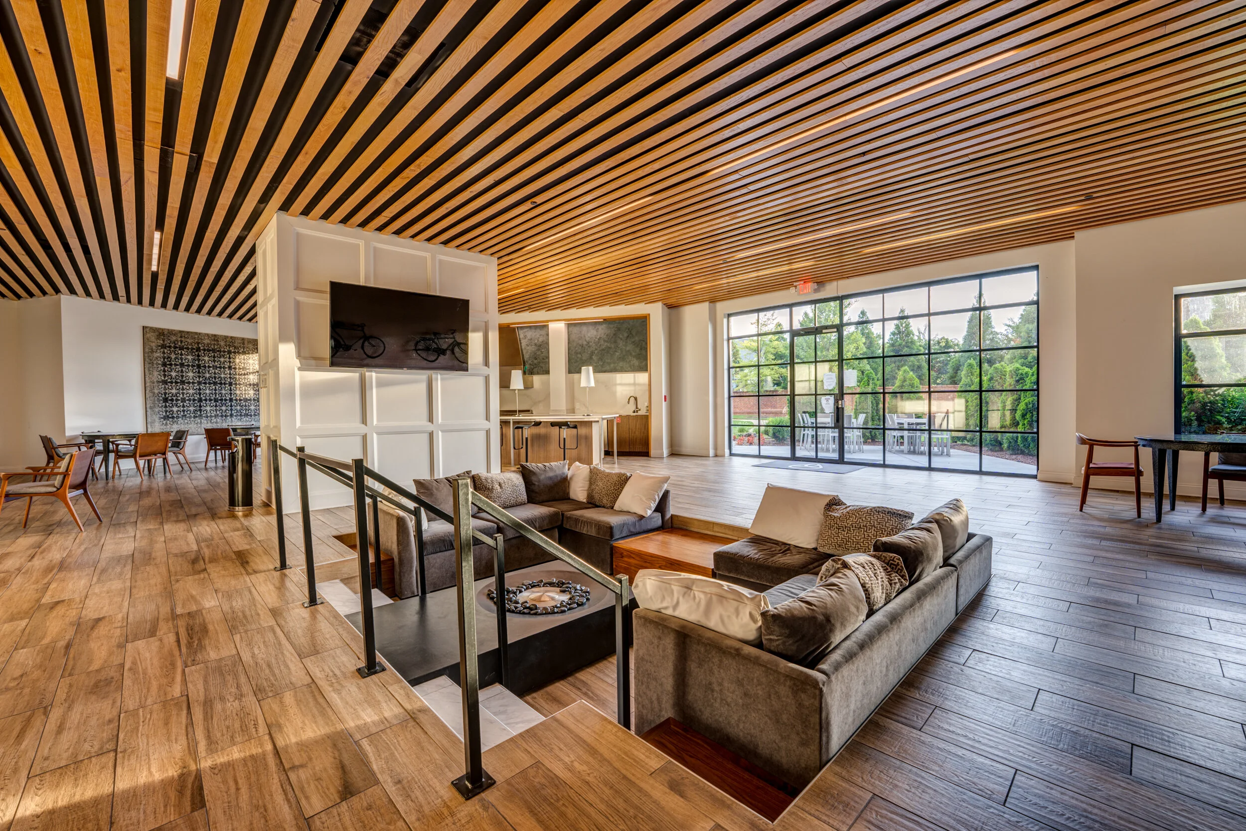 Spacious indoor lounge area with wooden ceiling and flooring, large glass windows, sectional sofas, and a television mounted on a partition wall.