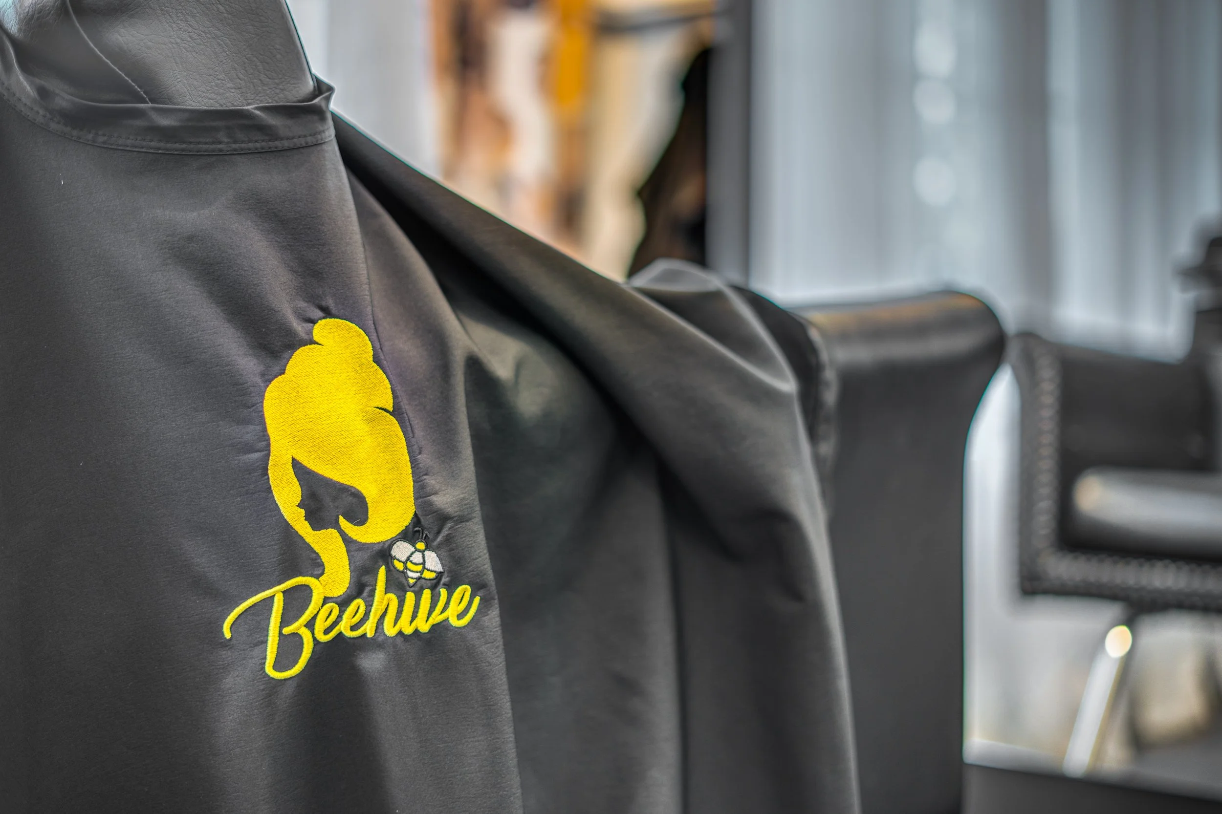 A black apron or jacket with a yellow embroidered logo of a beehive and the word "Beehive" on it, draped over a chair inside a room with natural light. Memphis Photography.