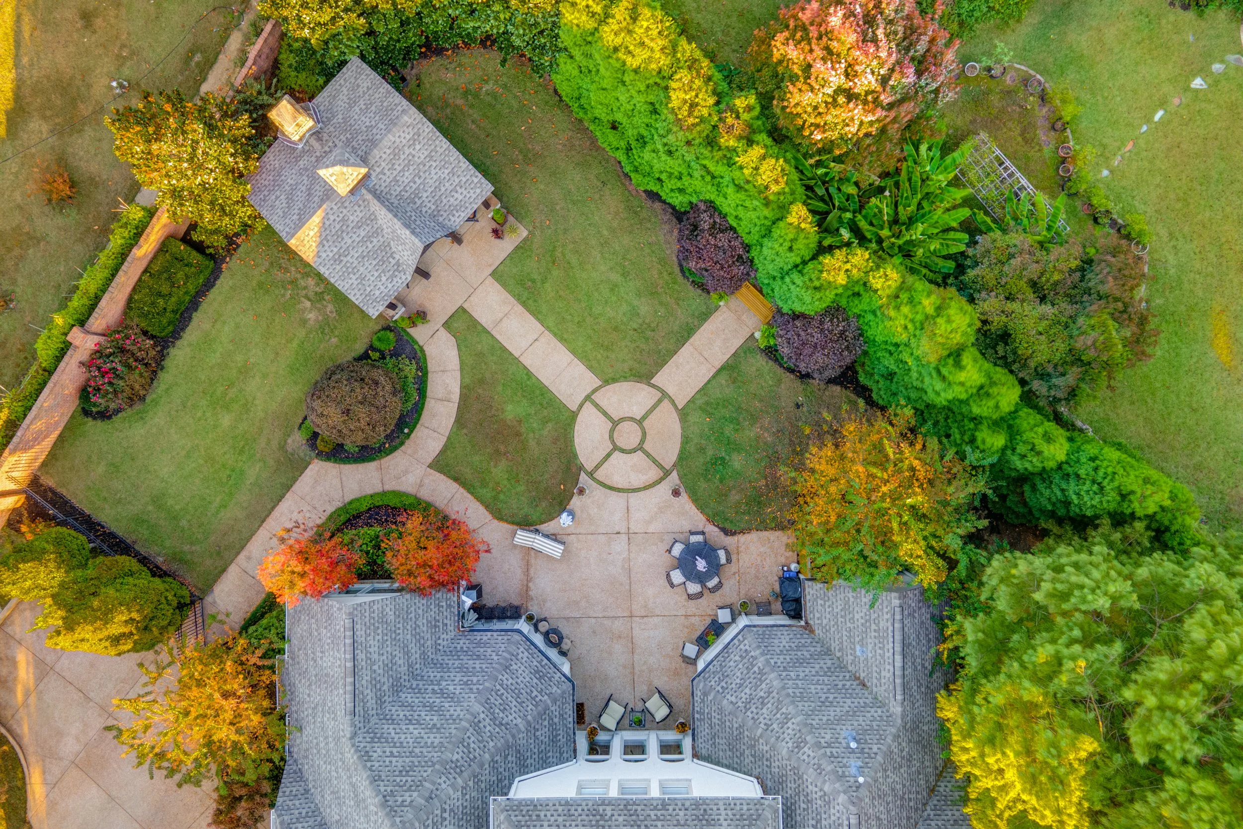 An aerial view of a backyard featuring a circular pathway, landscaped garden beds, mature trees with fall foliage, a patio with outdoor furniture, and a house with a gray shingled roof. Memphis real estate photography. 