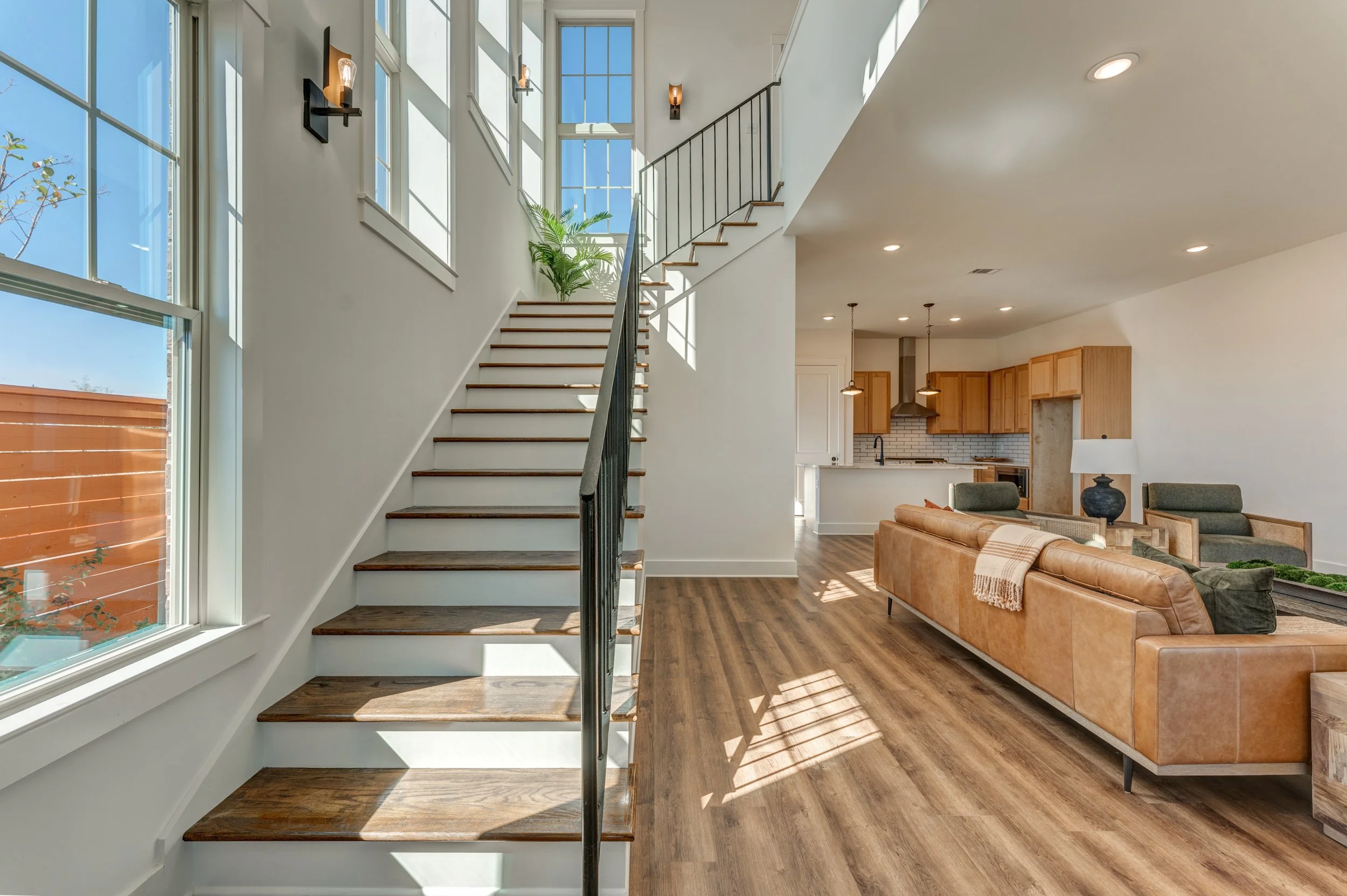 Modern living room with wooden flooring, large windows, a staircase with wooden steps and black railing leading upstairs, a brown leather sofa, green armchairs, a black table lamp, and a kitchen in the background with wooden cabinets, stainless steel