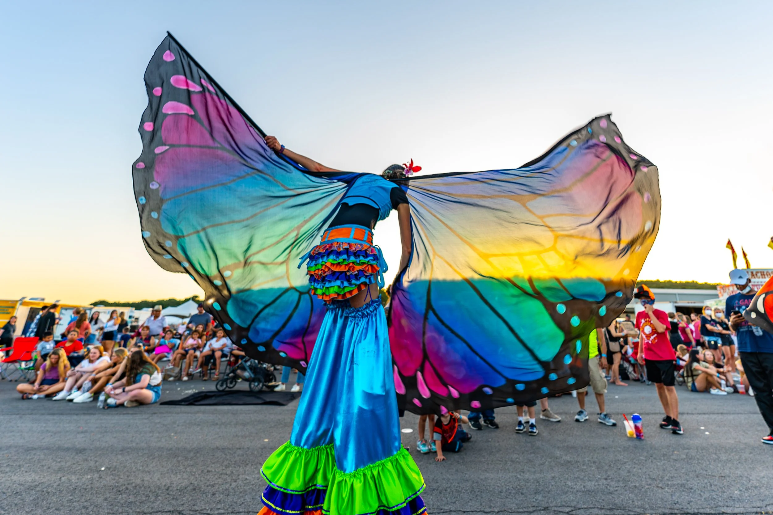 Performer in colorful butterfly costume with large rainbow wings at outdoor festival with seated crowd and tents in background during sunset.