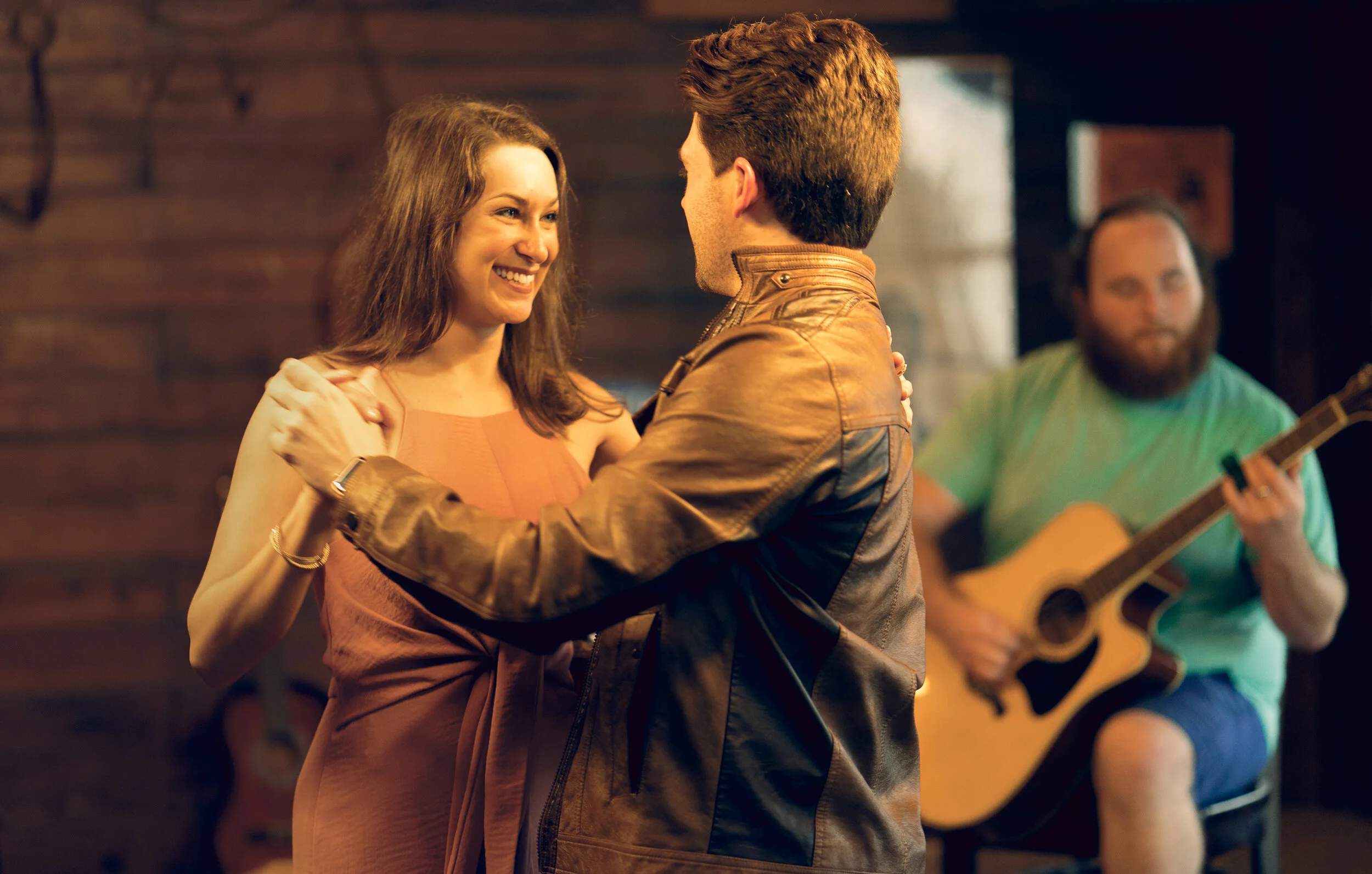 A young couple dancing closely together in a warmly lit room, with a musician playing guitar in the background. Memphis Photography. 