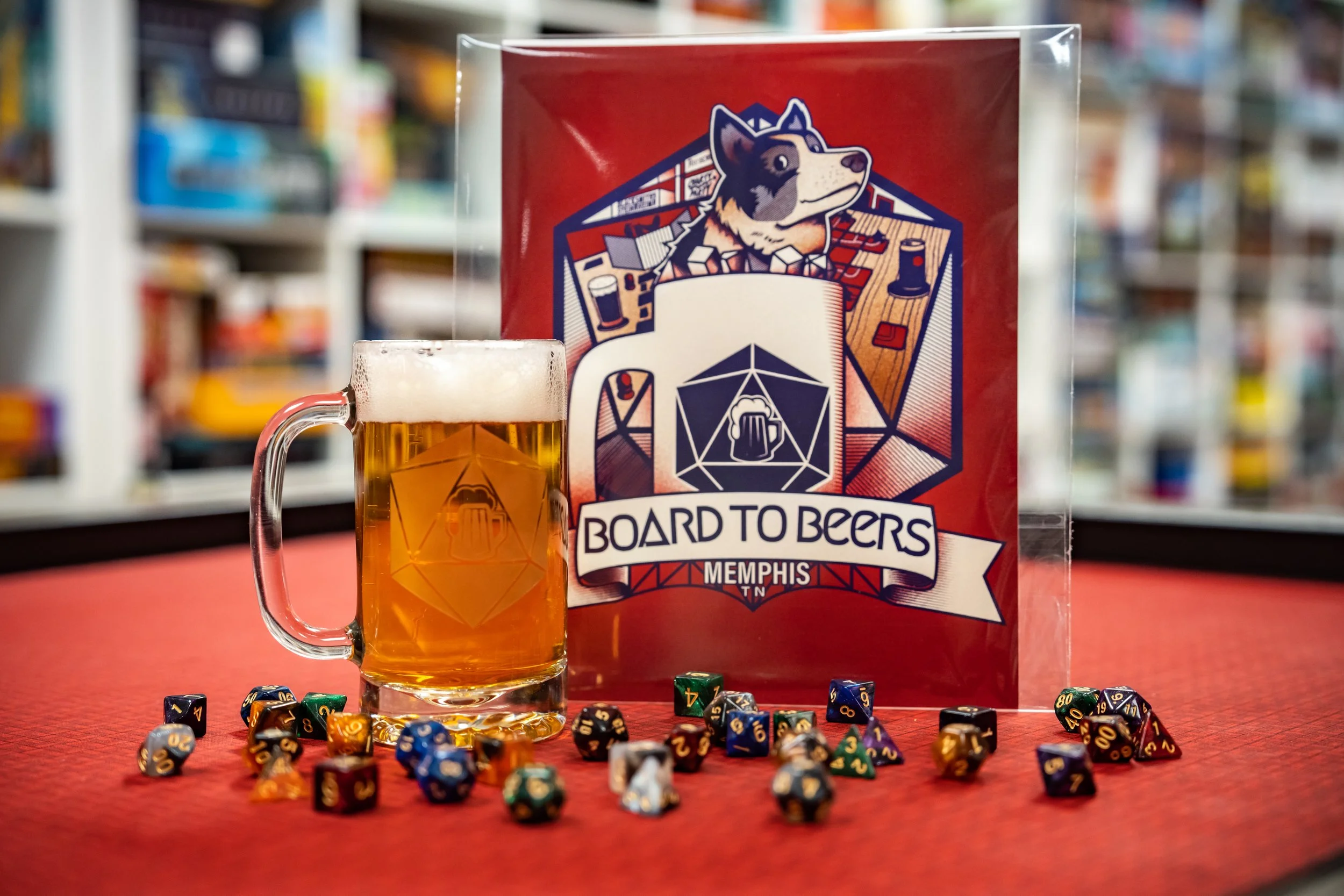 A beer mug with foam, decorated with a logo, on a red tabletop. Behind it is a sign or poster with a cartoon dog logo and the text 'Board to Beers Memphis TN'. Several colorful polyhedral dice are scattered in front of the mug and sign.