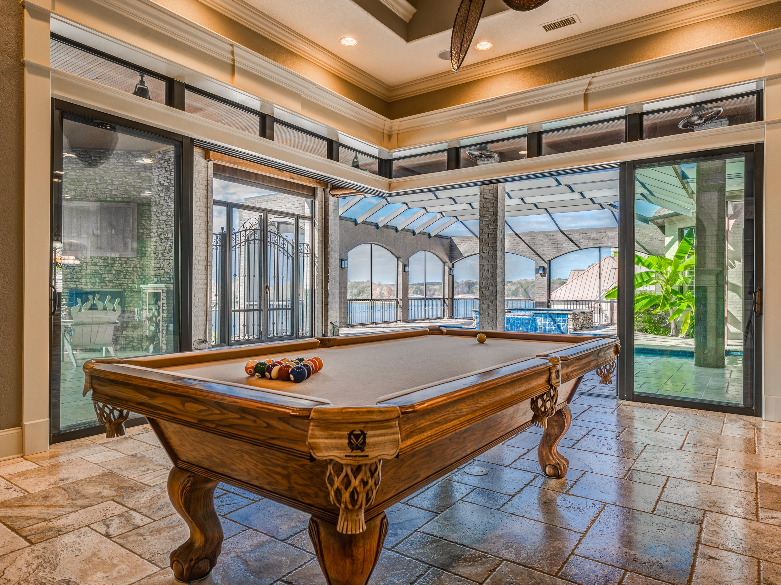 Indoor pool room with a wooden billiard table next to glass sliding doors leading to an outdoor patio with a pool and scenic view. Memphis real estate photography. 