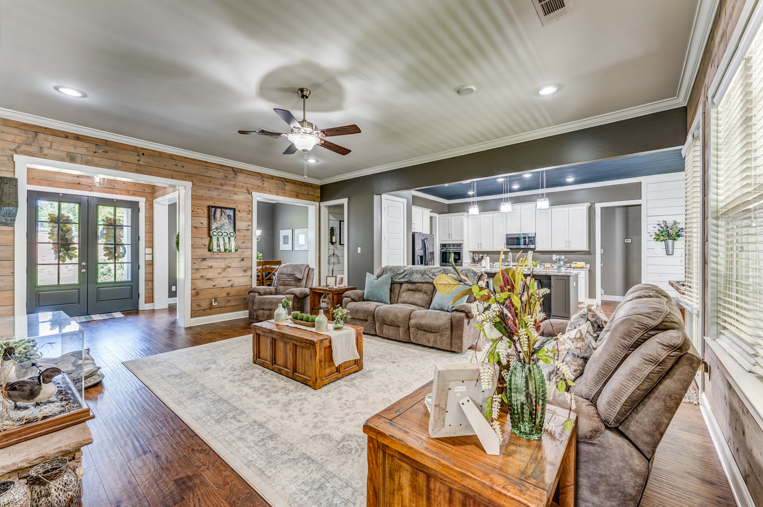 A spacious living room with wooden flooring, a beige couch, and wooden coffee table, decorated with plants, vases, and framed photos. The room opens into a modern white kitchen with pendant lighting. Memphis real estate photography. 