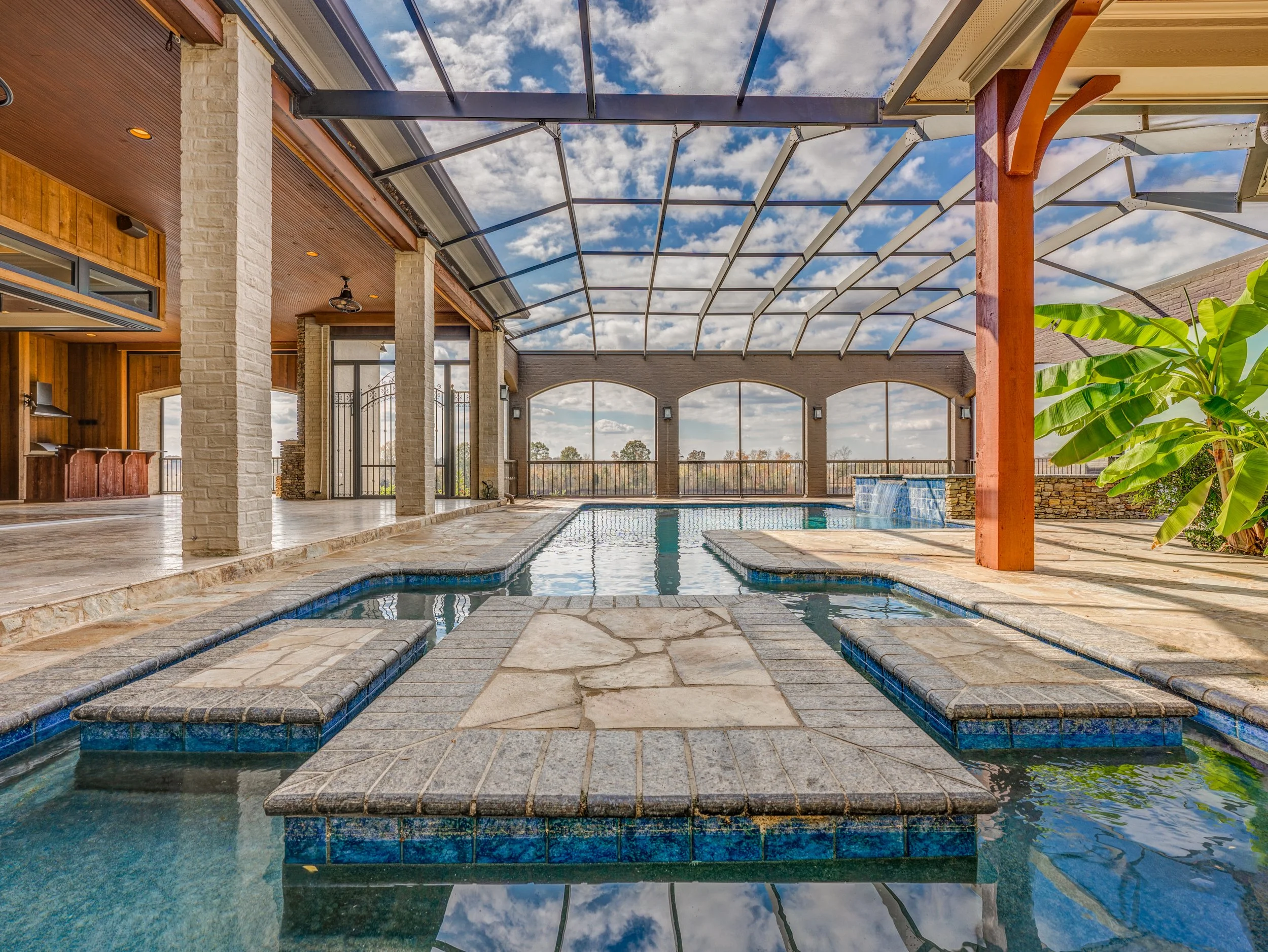 Indoor swimming pool area with a covered glass ceiling, stone and wood decor, and a view of the outside sky with clouds. There is a hot tub with built-in benches, a waterfall feature, and a large green plant. Memphis real estate photography. 