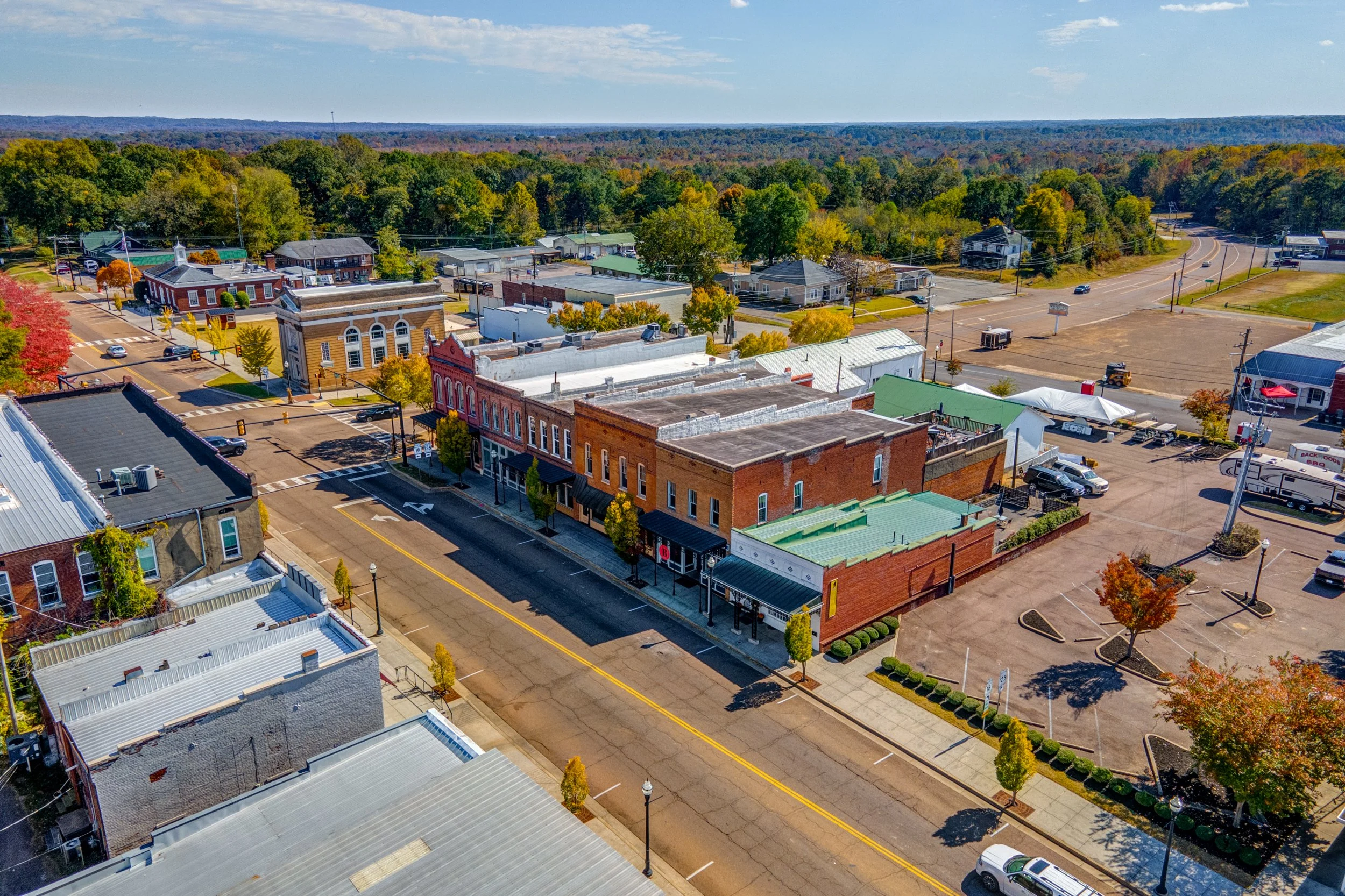 Aerial view of a small town with colorful brick buildings, a main street, trees with fall foliage, and a parking lot surrounded by shops and restaurants.
