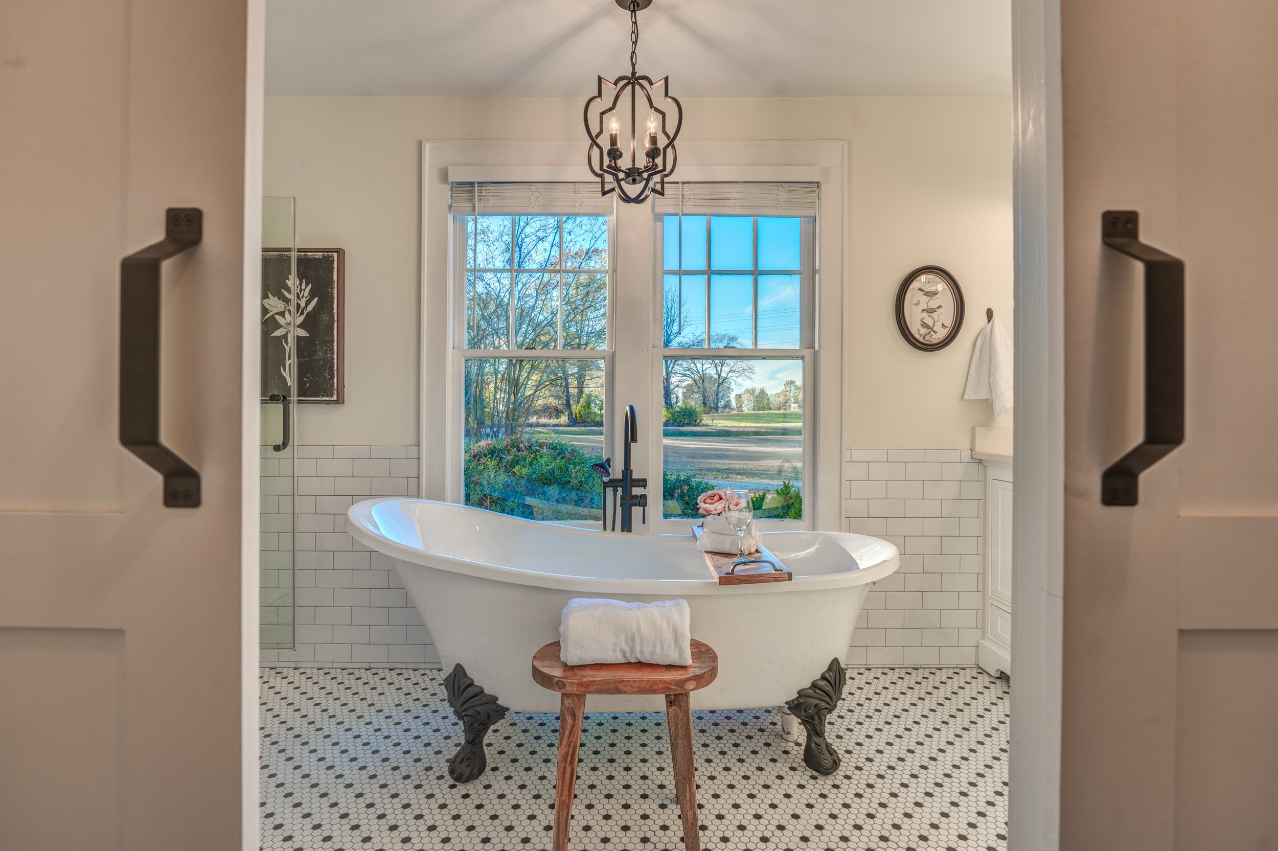 A bathroom with a classic clawfoot bathtub positioned in front of large windows showing an outdoor view, with a black light fixture hanging from the ceiling, and white subway tile halfway up the wall. Memphis real estate photography. 