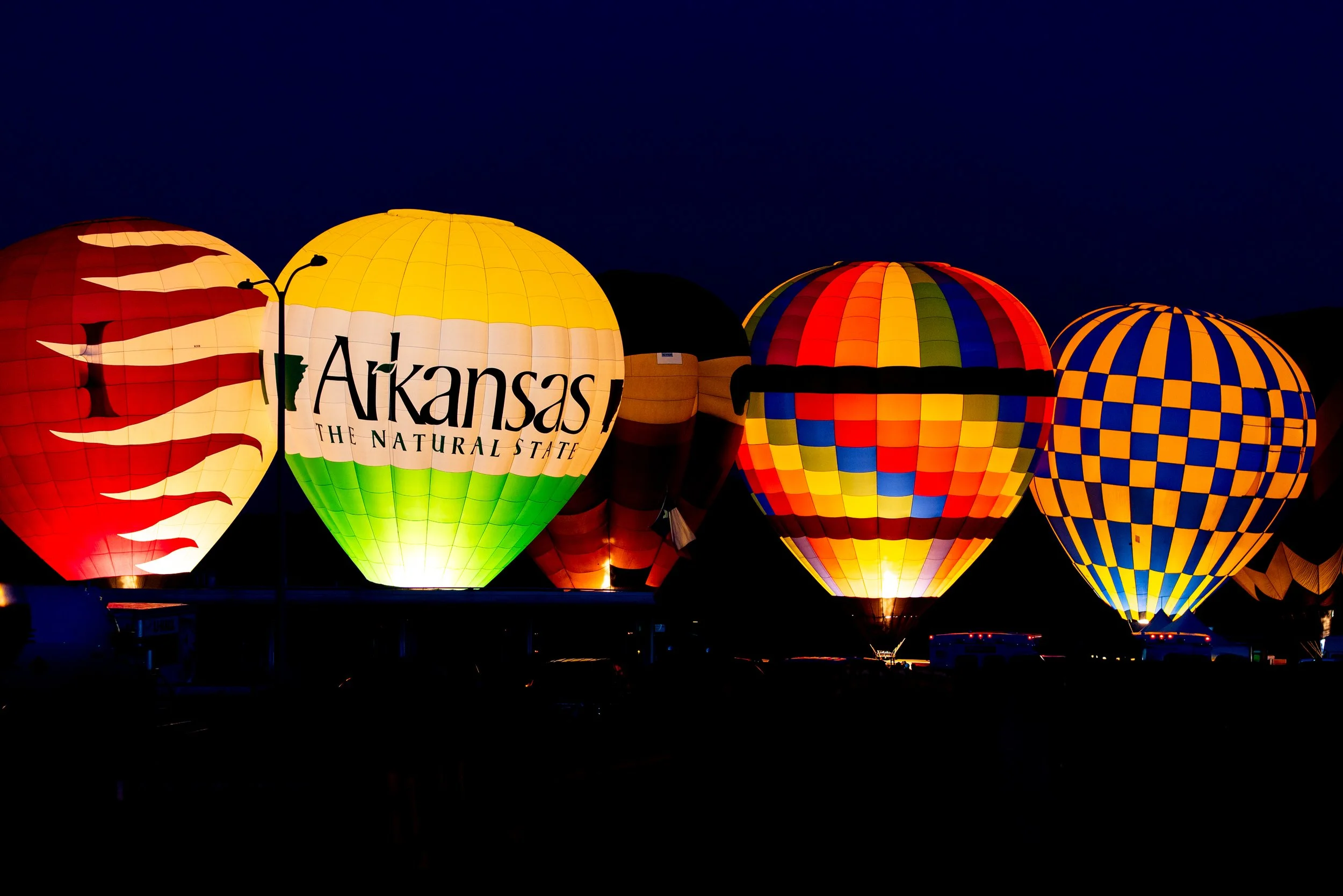 Colorful hot air balloons illuminated at night, including one with Arkansas State and The Natural State written on it, and others with checkered and striped patterns.