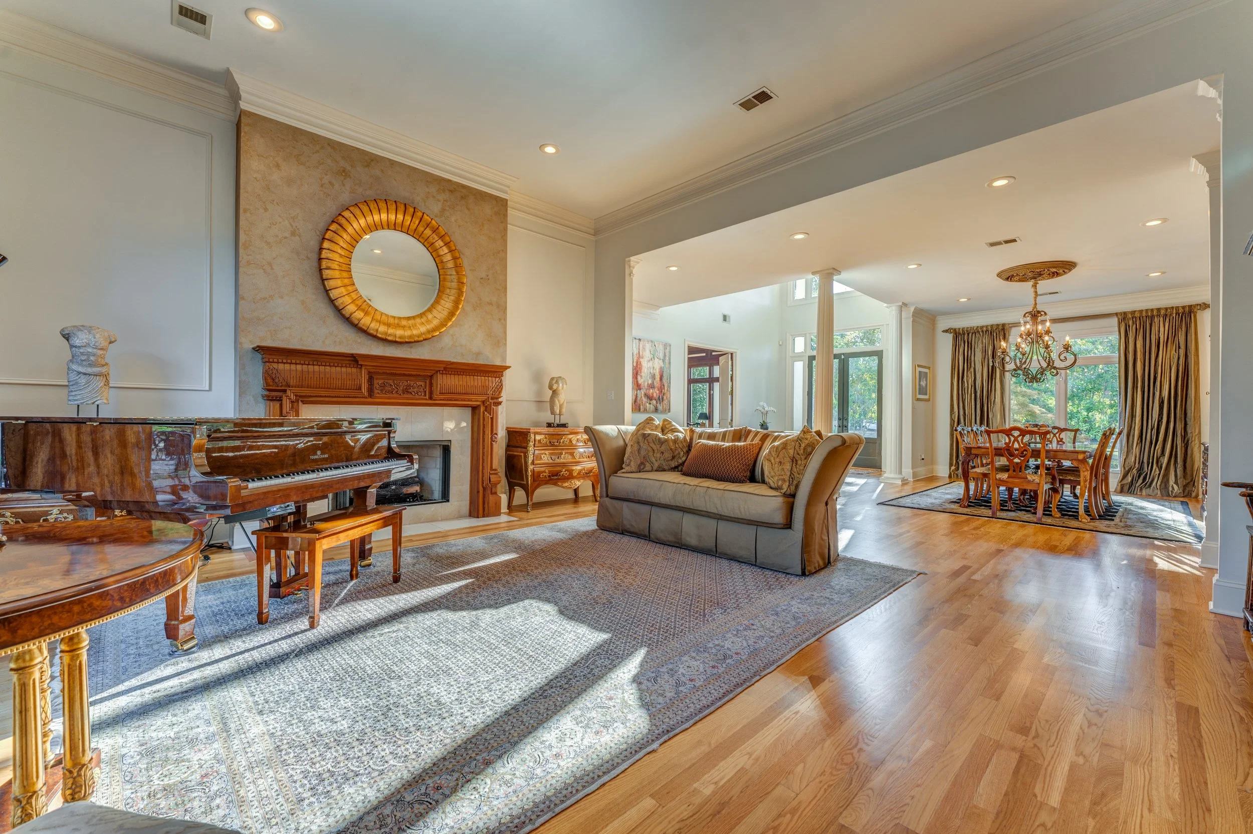 Living room with hardwood floors, a piano, a fireplace with wooden mantel, a beige sofa with decorative pillows, a patterned rug, and a dining area with a chandelier and large windows with curtains. Memphis real estate photography. 