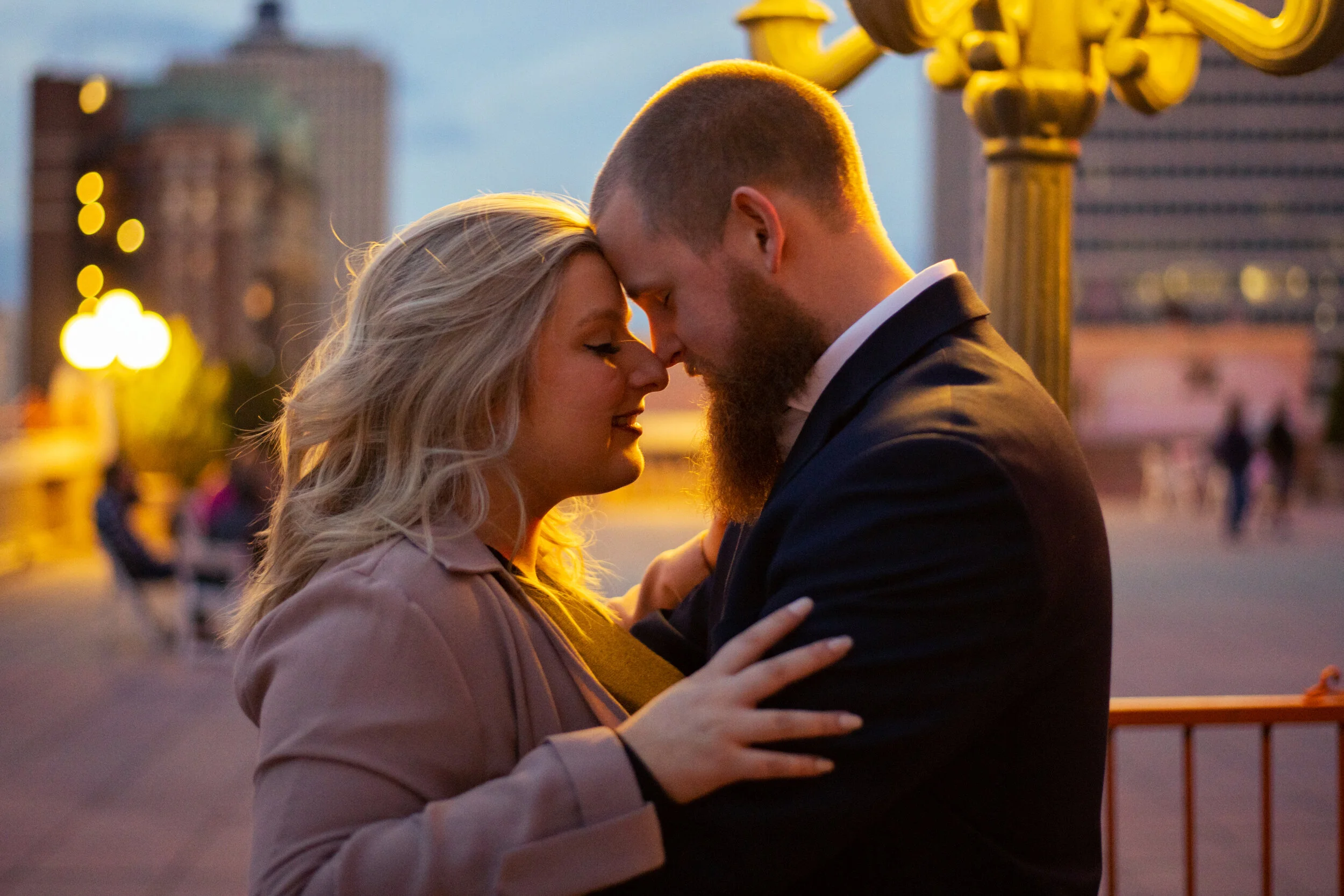 A couple with their foreheads touching in an intimate moment during sunset, with the city skyline and blurred lights in the background.