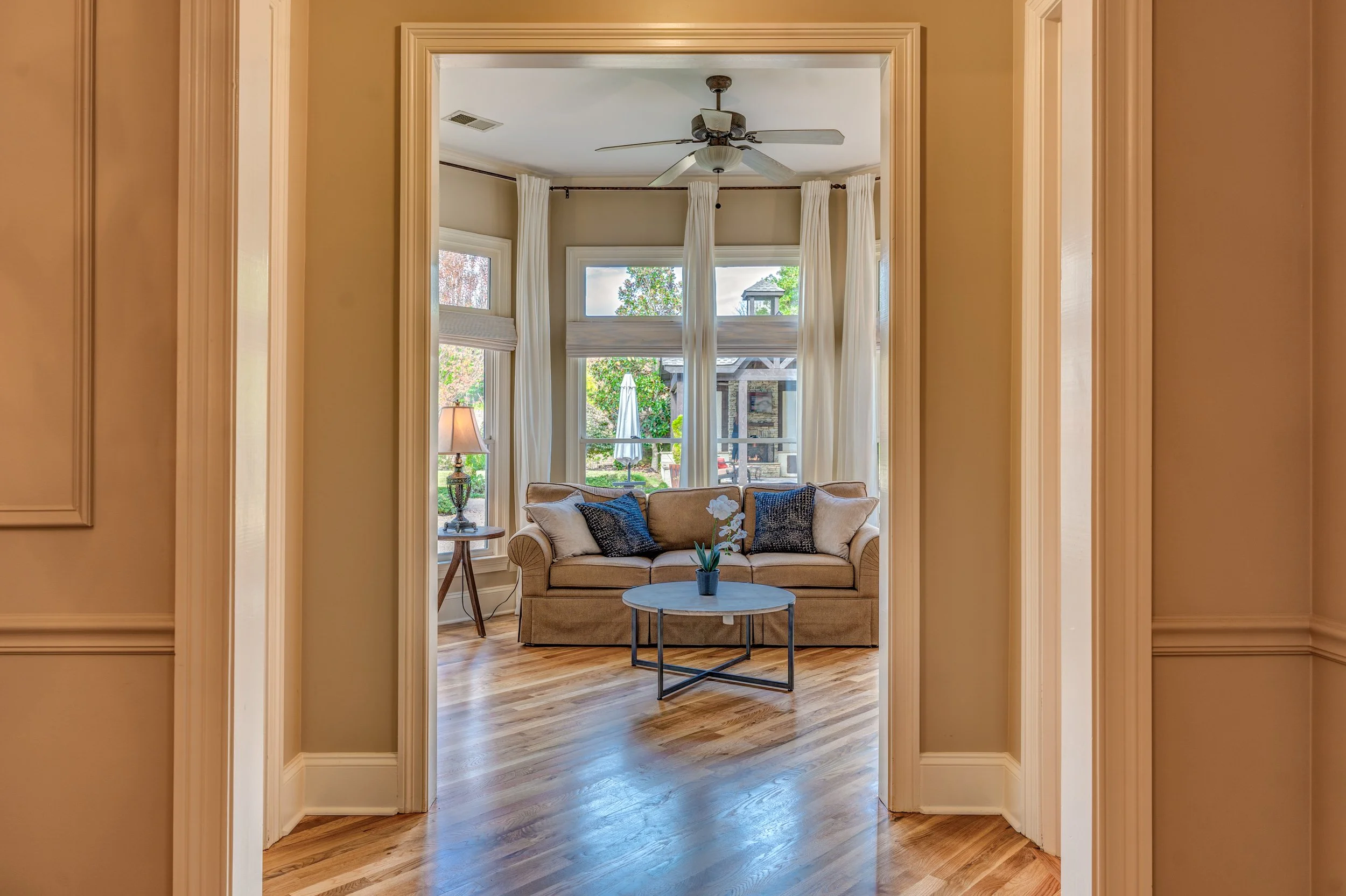 Living room with beige sofa, throw pillows, small round coffee table, lamp, and large windows with white curtains, overlooking a backyard patio. Memphis real estate photography. 