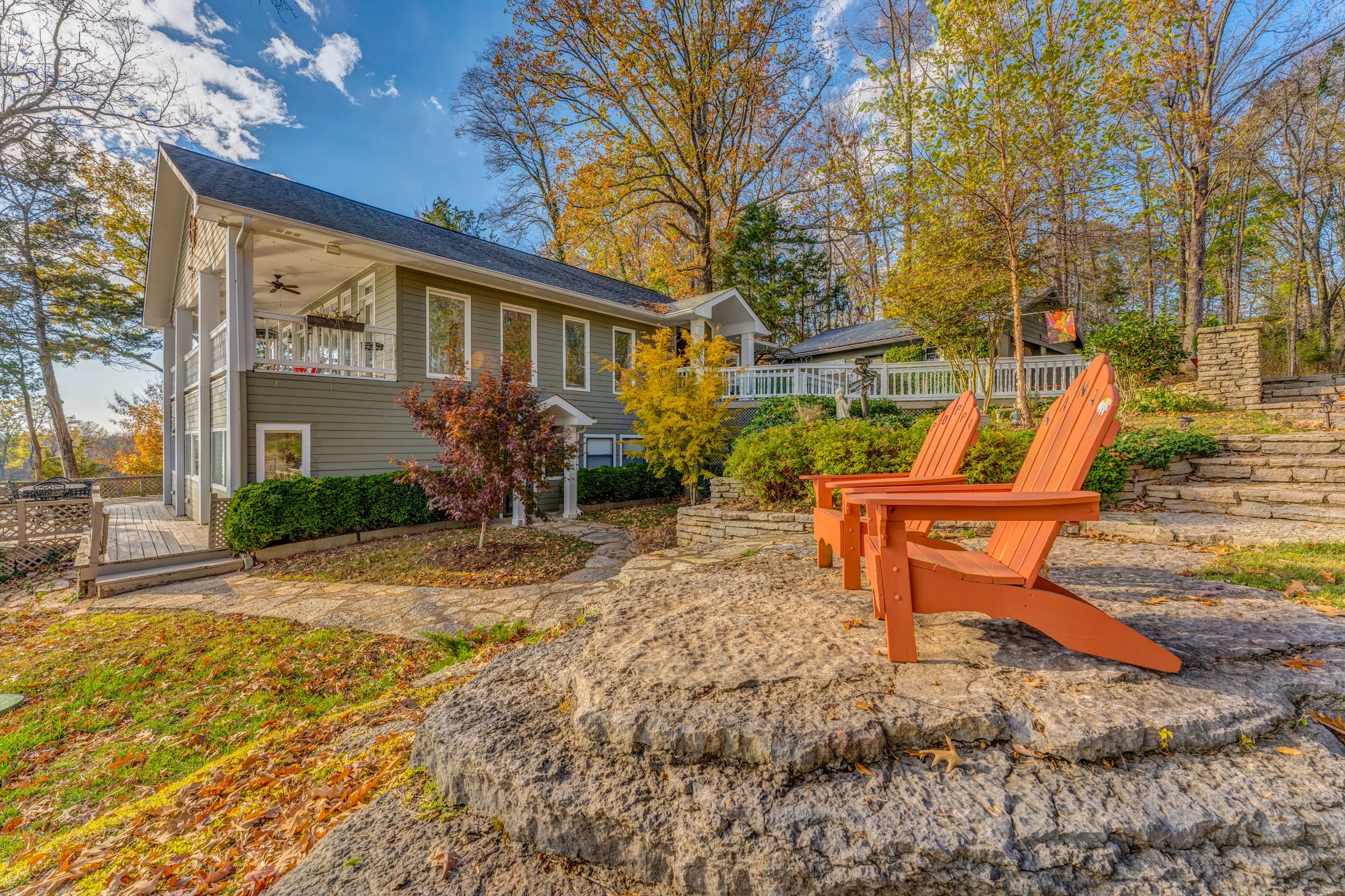 Colorful autumn backyard scene with two orange Adirondack chairs on large rock, surrounded by trees in fall foliage, a modern house with a deck, and a partly cloudy sky. Memphis real estate photography. 