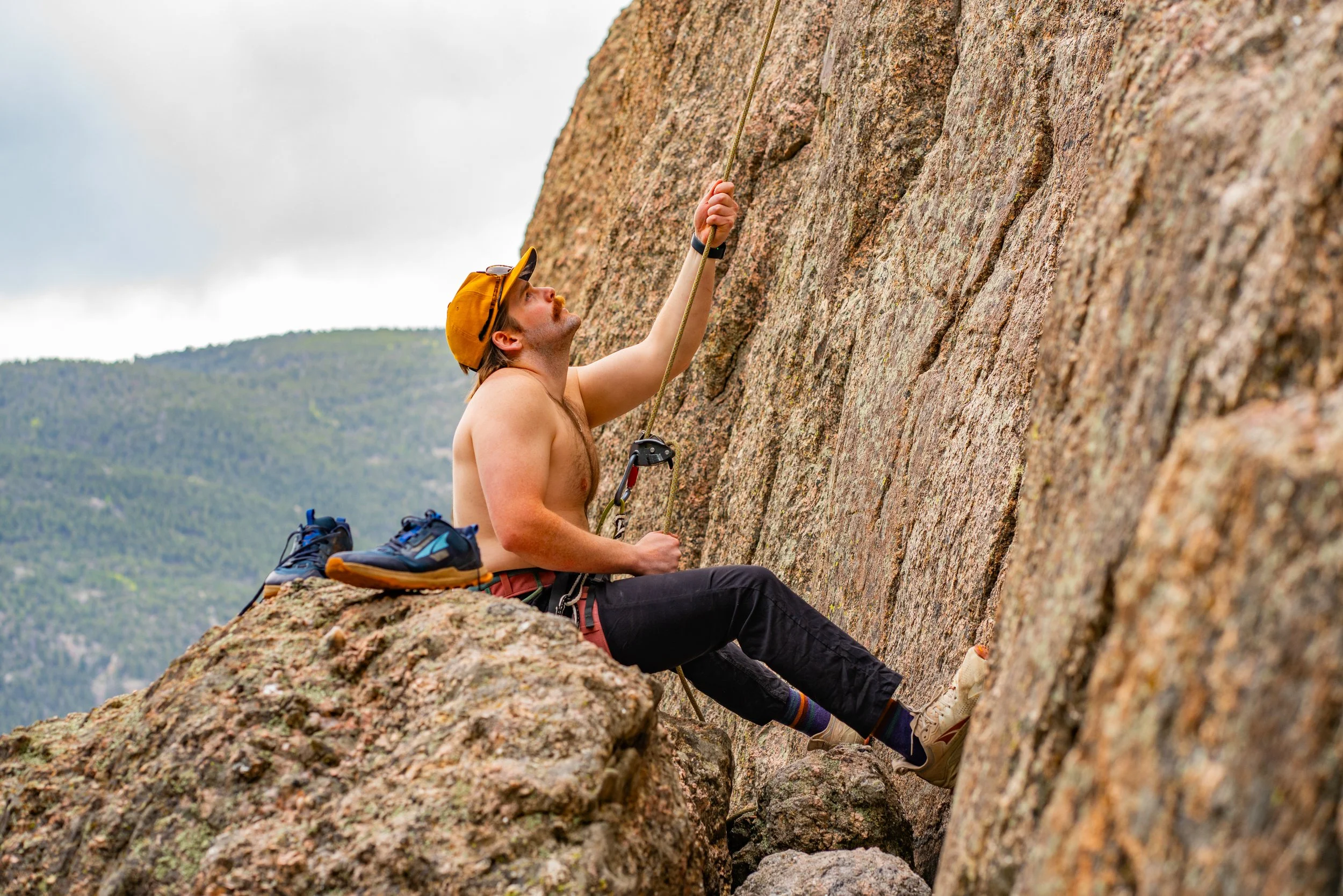 A shirtless man wearing a yellow cap, black pants, and climbing shoes is rock climbing on a steep granite wall outdoors. He is seated on a large rock with climbing gear beside him, reaching with his right hand for a hold on the wall while holding a c