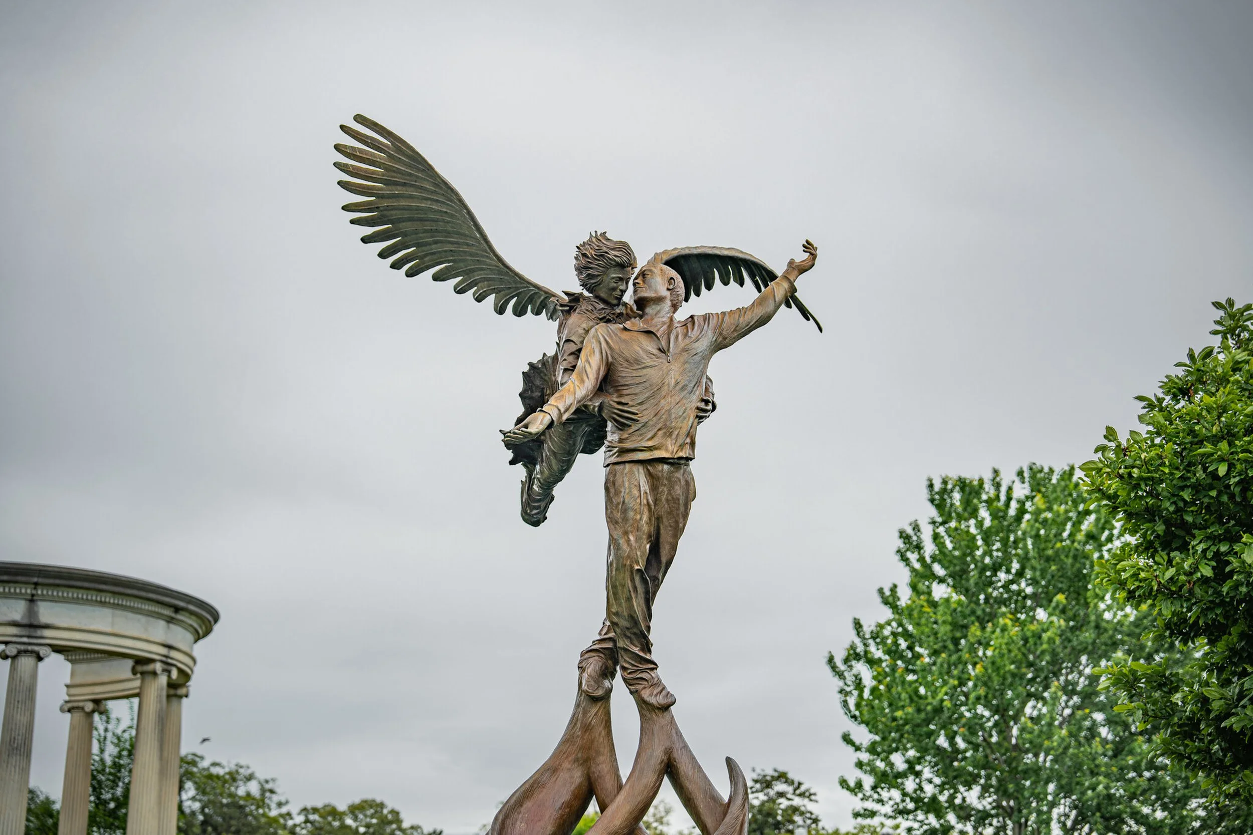 Bronze sculpture of a man with arms outstretched, carrying a winged figure on his back, set against a cloudy sky and surrounded by green trees. Memphis architectural photography.