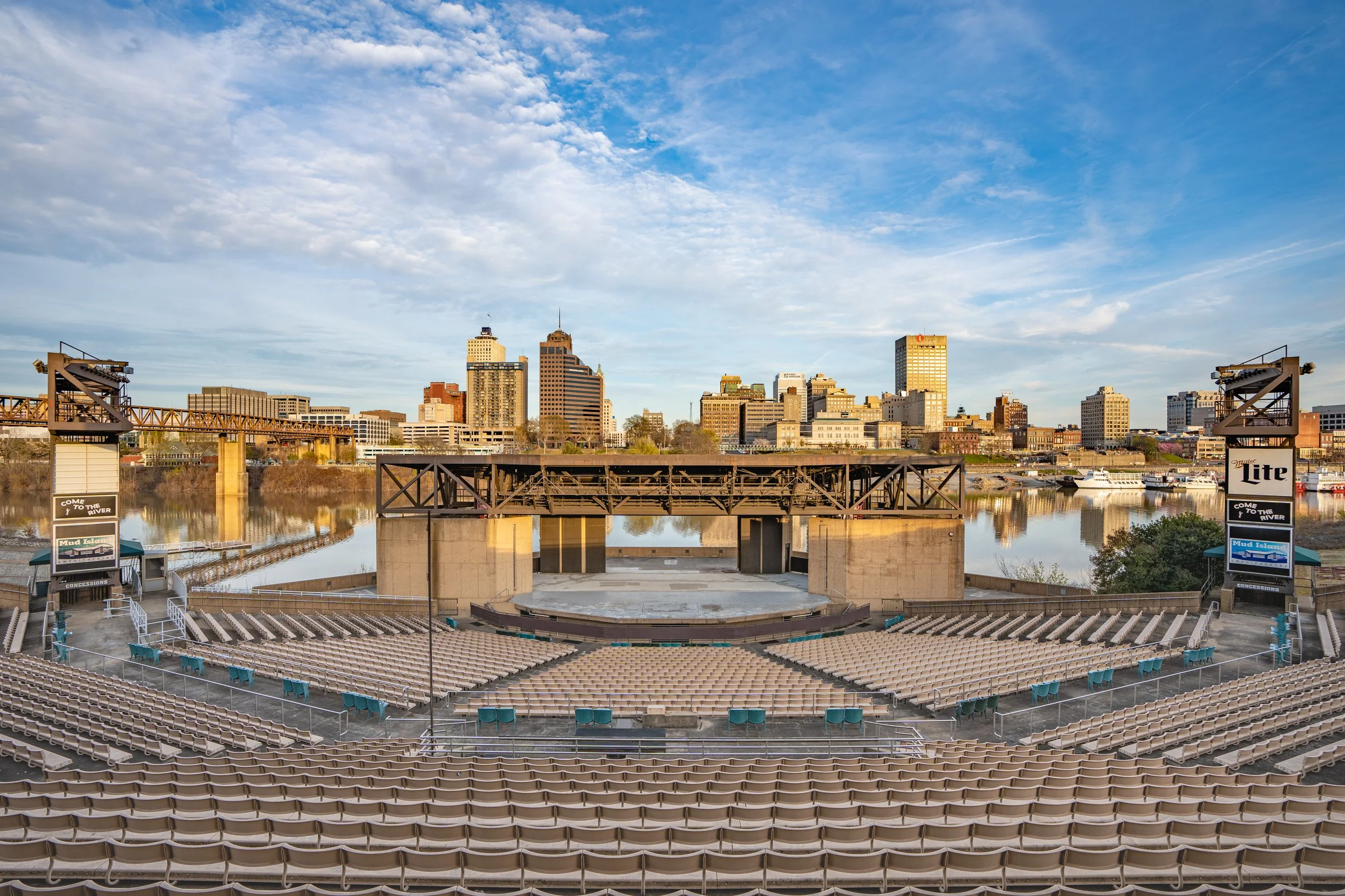 An outdoor amphitheater with rows of seats facing a stage, overlooking a river with a city skyline in the background under a partly cloudy sky. Memphis architectural photography.