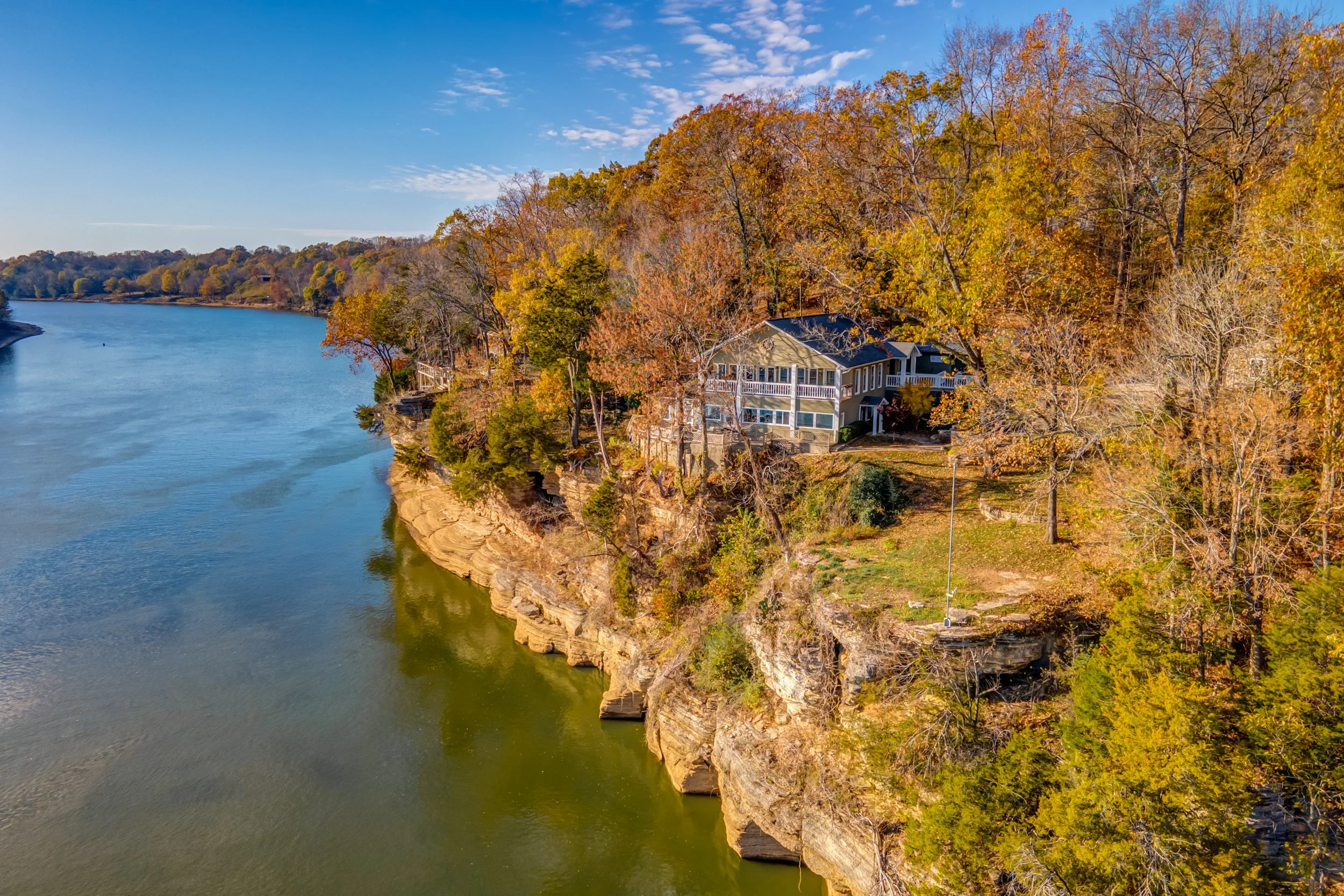 Aerial view of a house on a cliff overlooking a river with fall foliage on trees.
