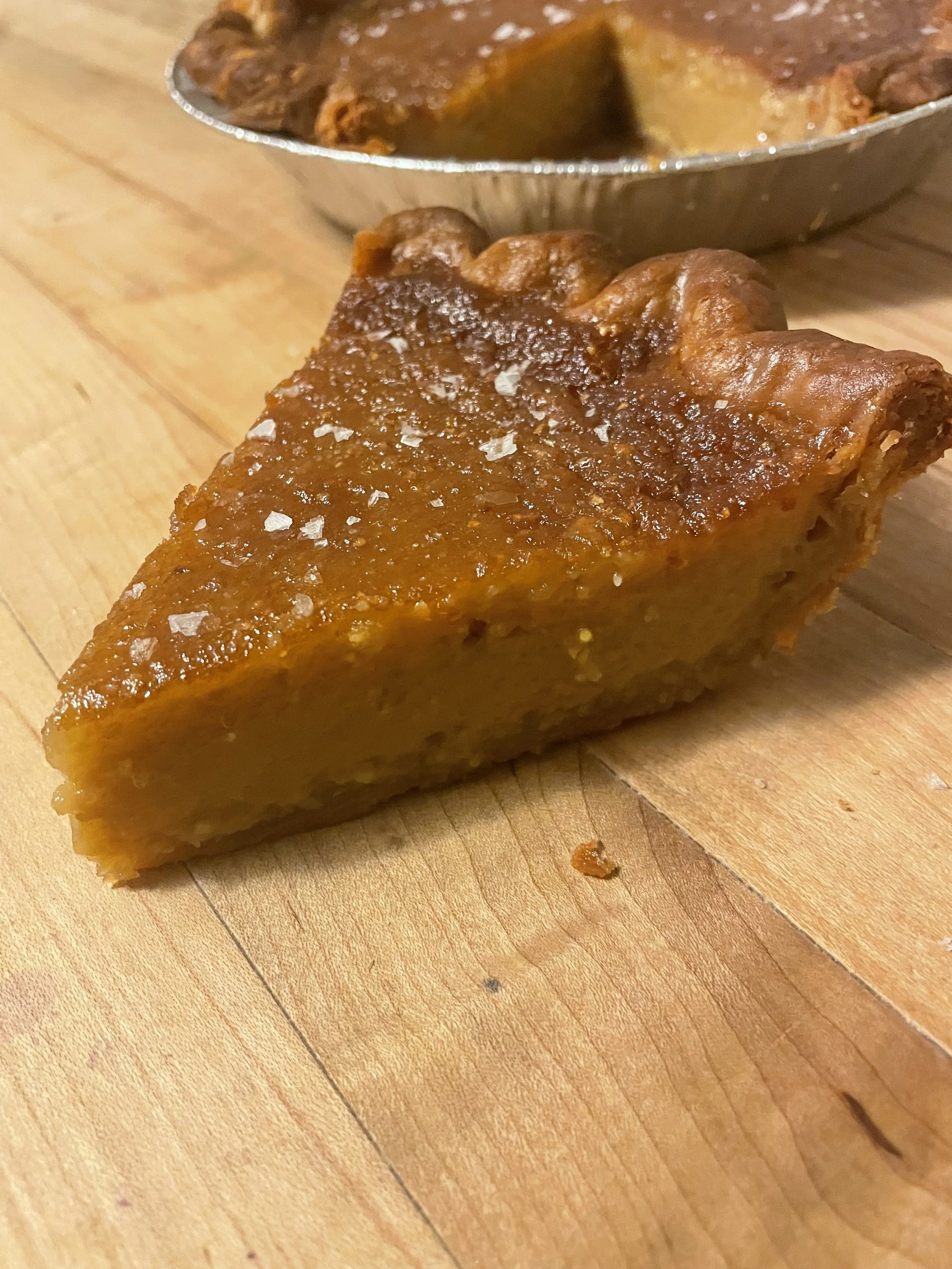 A slice of salted maple pie with a flaky crust, topped with salt, on a wooden surface with a pie dish in the background.