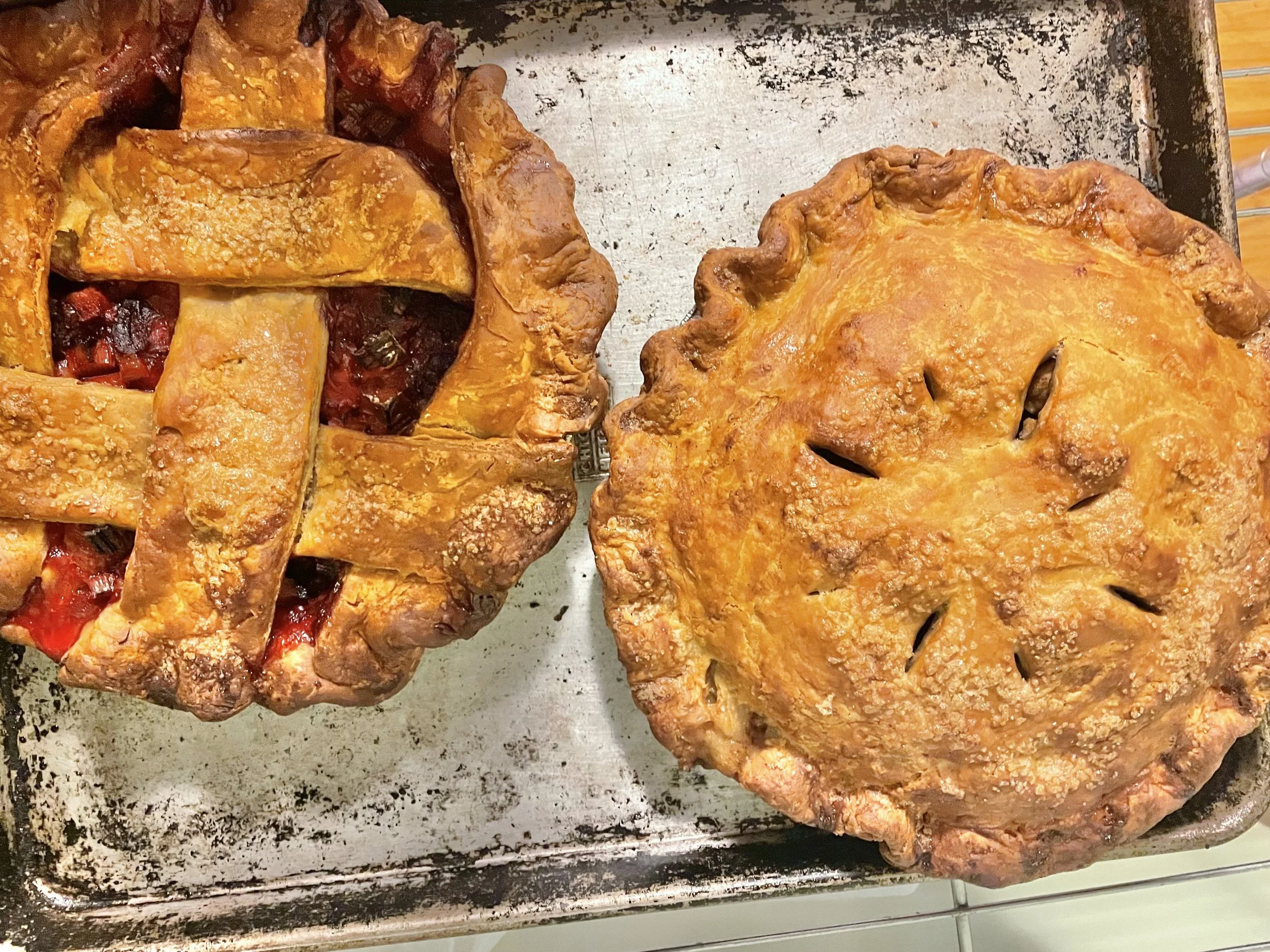 Two baked pies on a metal tray, one on the left with a lattice crust and filling spilling out, the other on the right with a crust and small slits on top.
