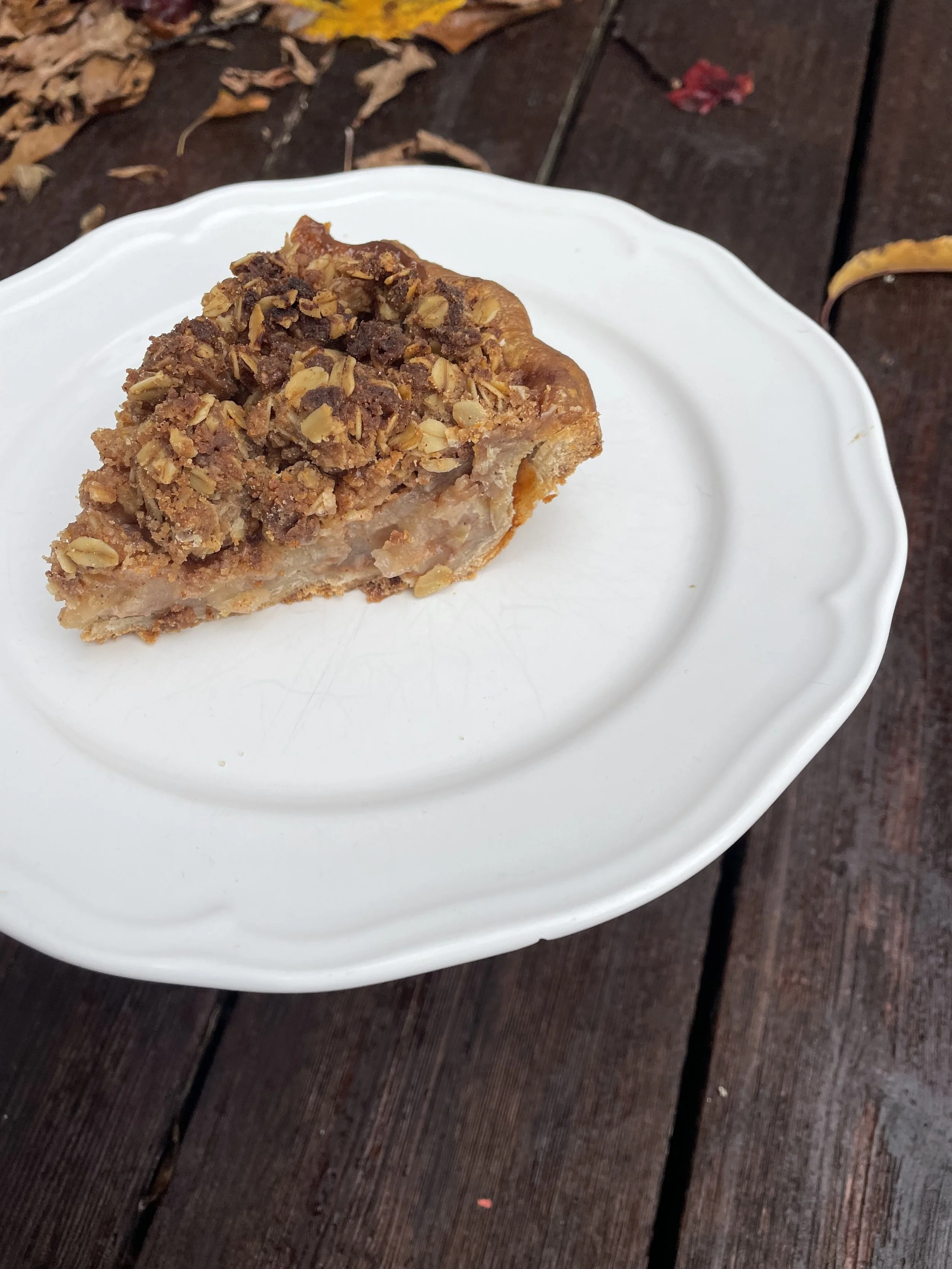 A slice of apple crumble pie on a white plate with a wooden surface and fallen autumn leaves in the background.