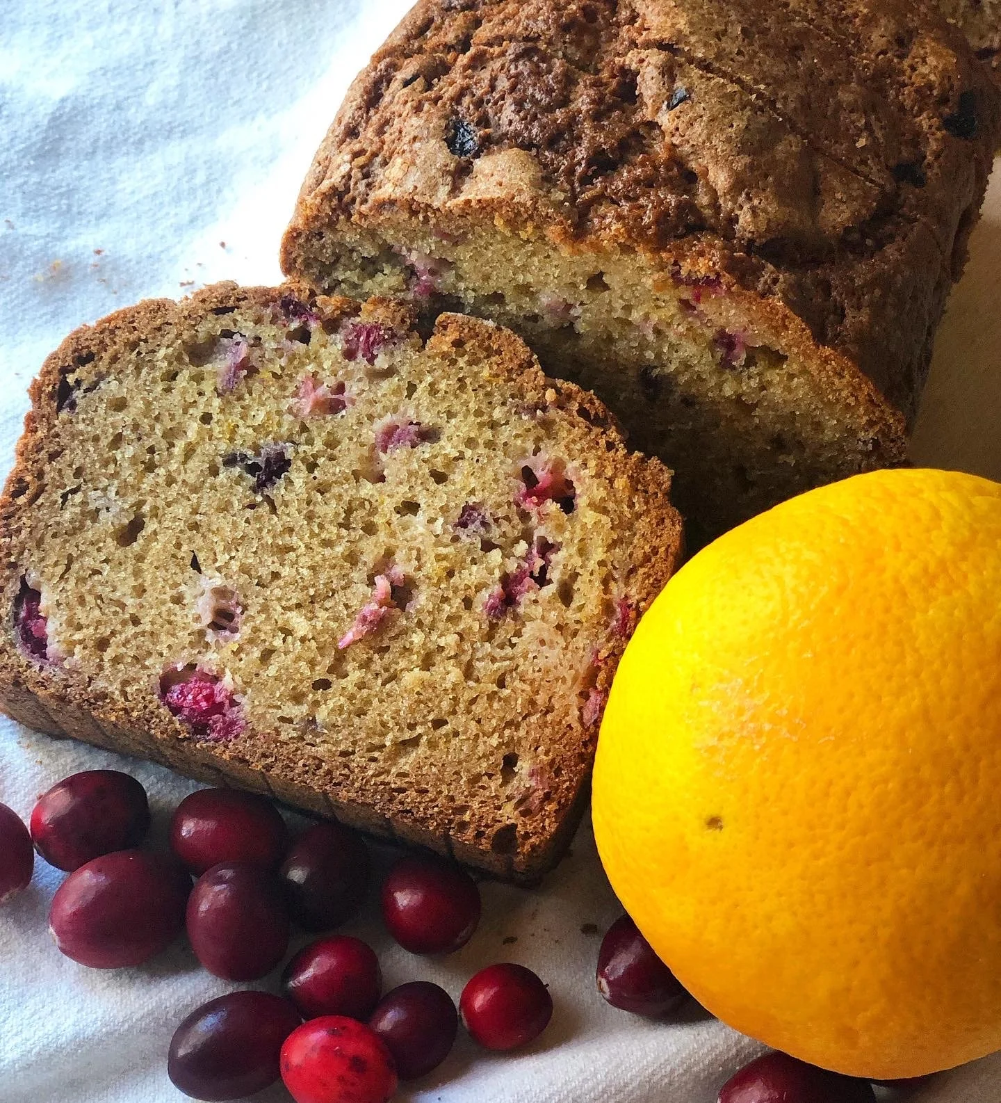 Slices of cranberry orange bread with fresh cranberries and a whole orange on a white surface.