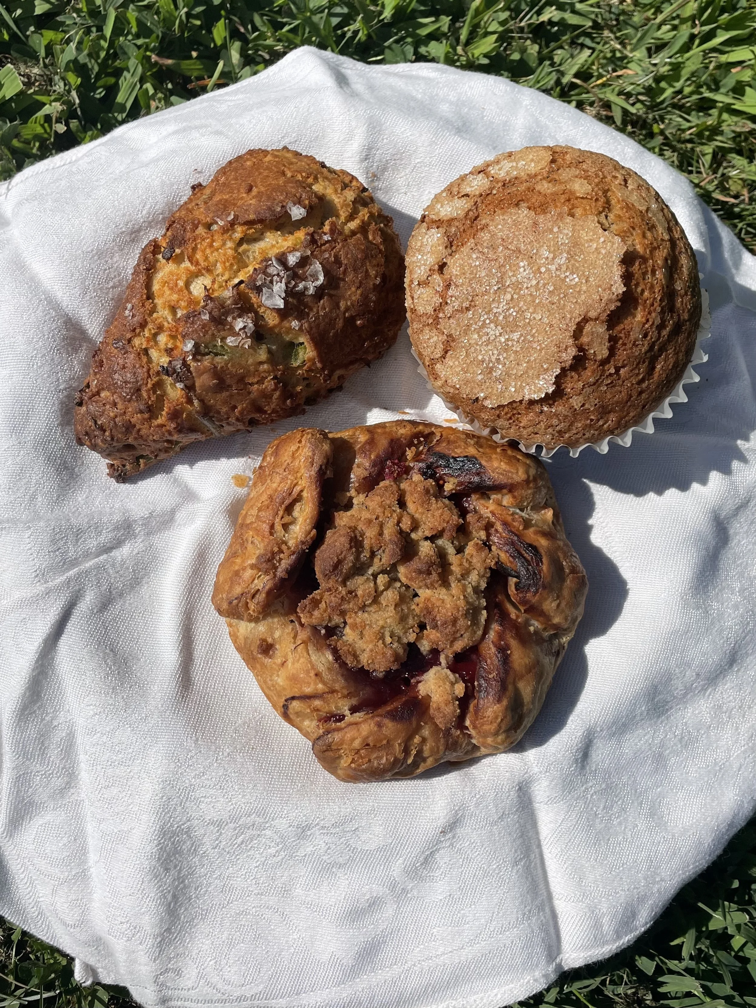 Three baked goods on white cloth, with green grass in background: a muffin with sugar topping, a galette with a crumbly topping, and a scone with sugar.