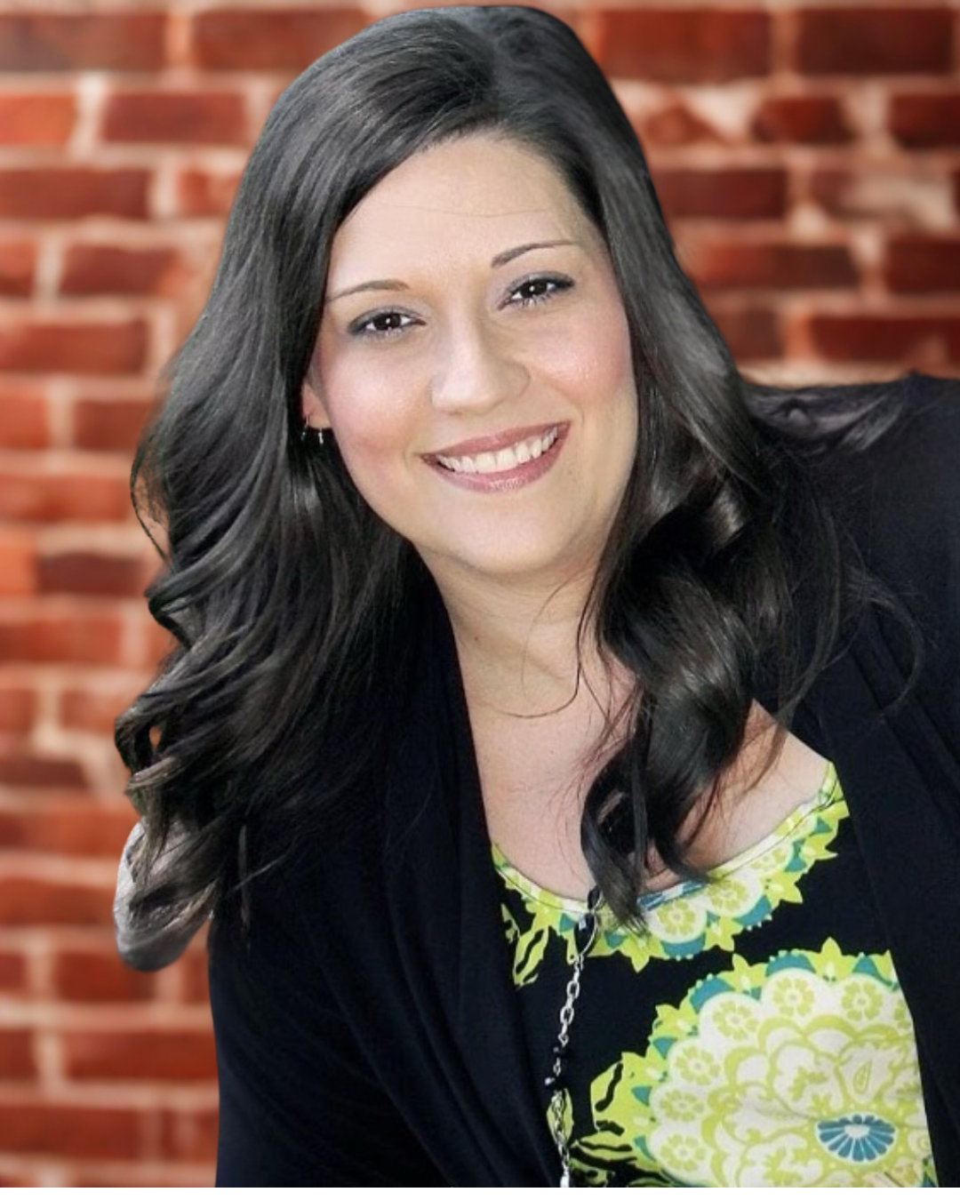 A woman with long dark wavy hair smiling at the camera, wearing a black blazer over a colorful yellow and green patterned top, with a brick wall in the background.