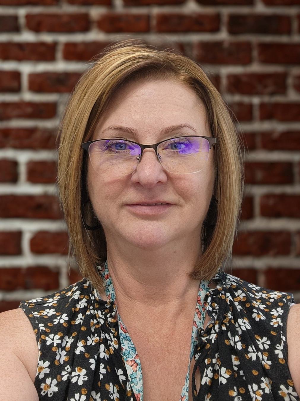 A woman with shoulder-length light brown hair wearing glasses and a sleeveless black top with a floral pattern, standing in front of a red brick wall.