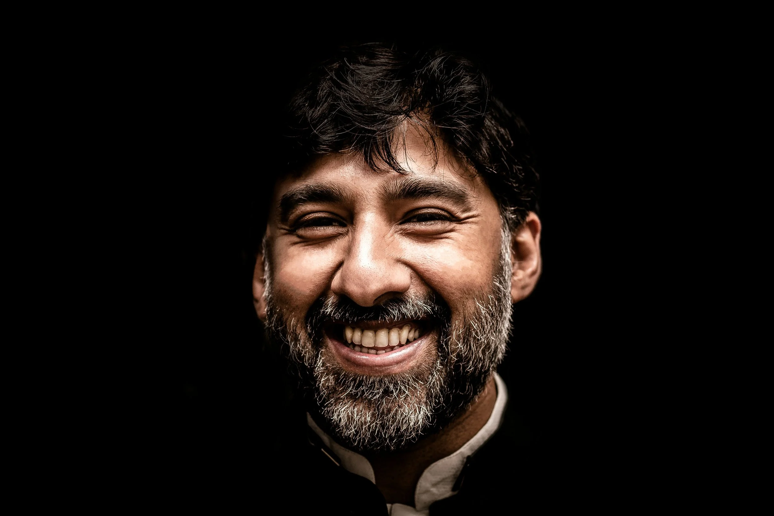 Close-up of a smiling middle-aged man with a beard, dark hair, and wearing a black suit against a black background.