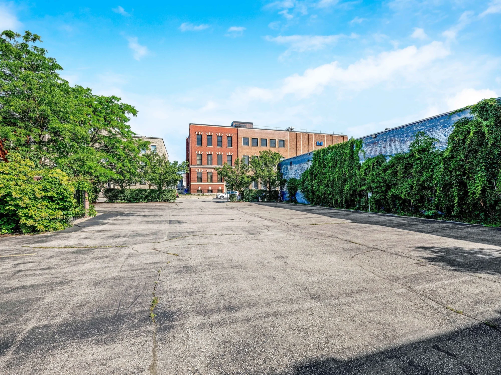 Wide view of the rear parking lot with mature trees and a brick building beyond