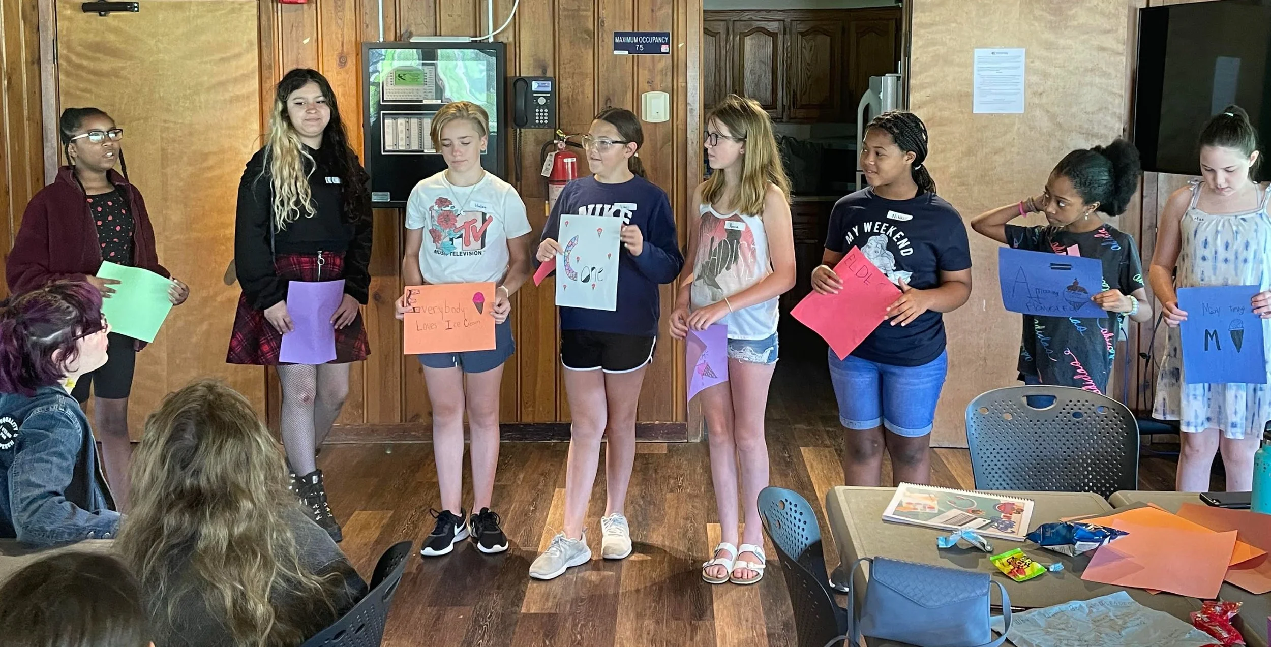 Group of young girls standing in a line, holding colorful signs, likely presenting or performing in a classroom or community room setting.