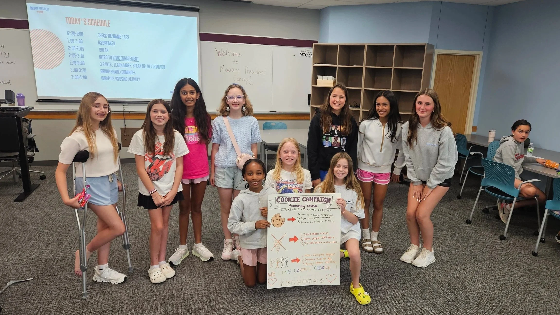 Group of young girls in a classroom, some holding a poster about a cookie campaign. The classroom has a whiteboard, a projector screen displaying the schedule, and some students sitting at desks to the side.
