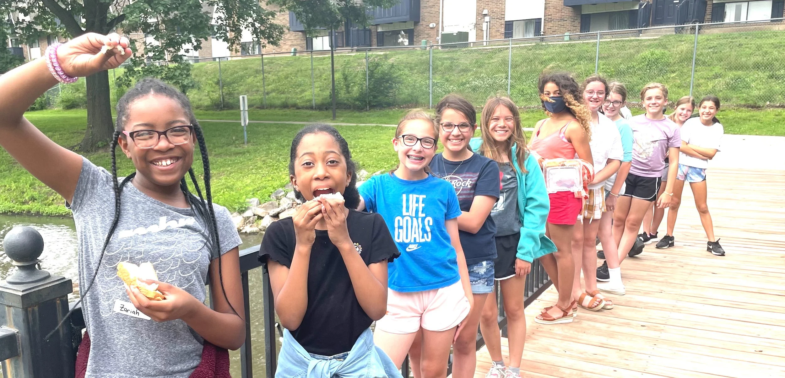 A group of young girls standing in a line on a wooden bridge outdoors, smiling and enjoying snacks, with a grassy park and buildings in the background.
