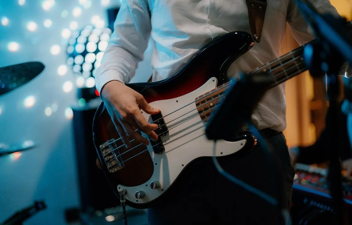A group of musicians performing at an indoor wedding, including a drummer, a woman playing tenor saxophone, a man playing trumpet, and a man playing alto saxophone, with a warm-toned textured wall in the background.