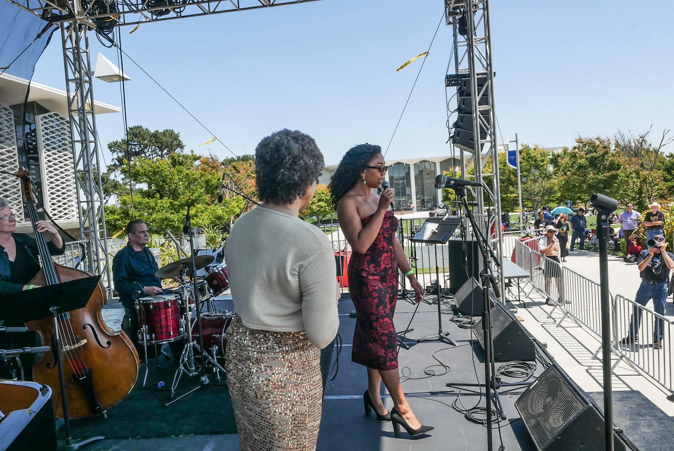 A woman in a strapless dress and high heels is speaking into a microphone on a stage during an outdoor event. She is accompanied by another woman in a beige top and leopard print skirt, and a band with a bassist and drummer is performing behind them.