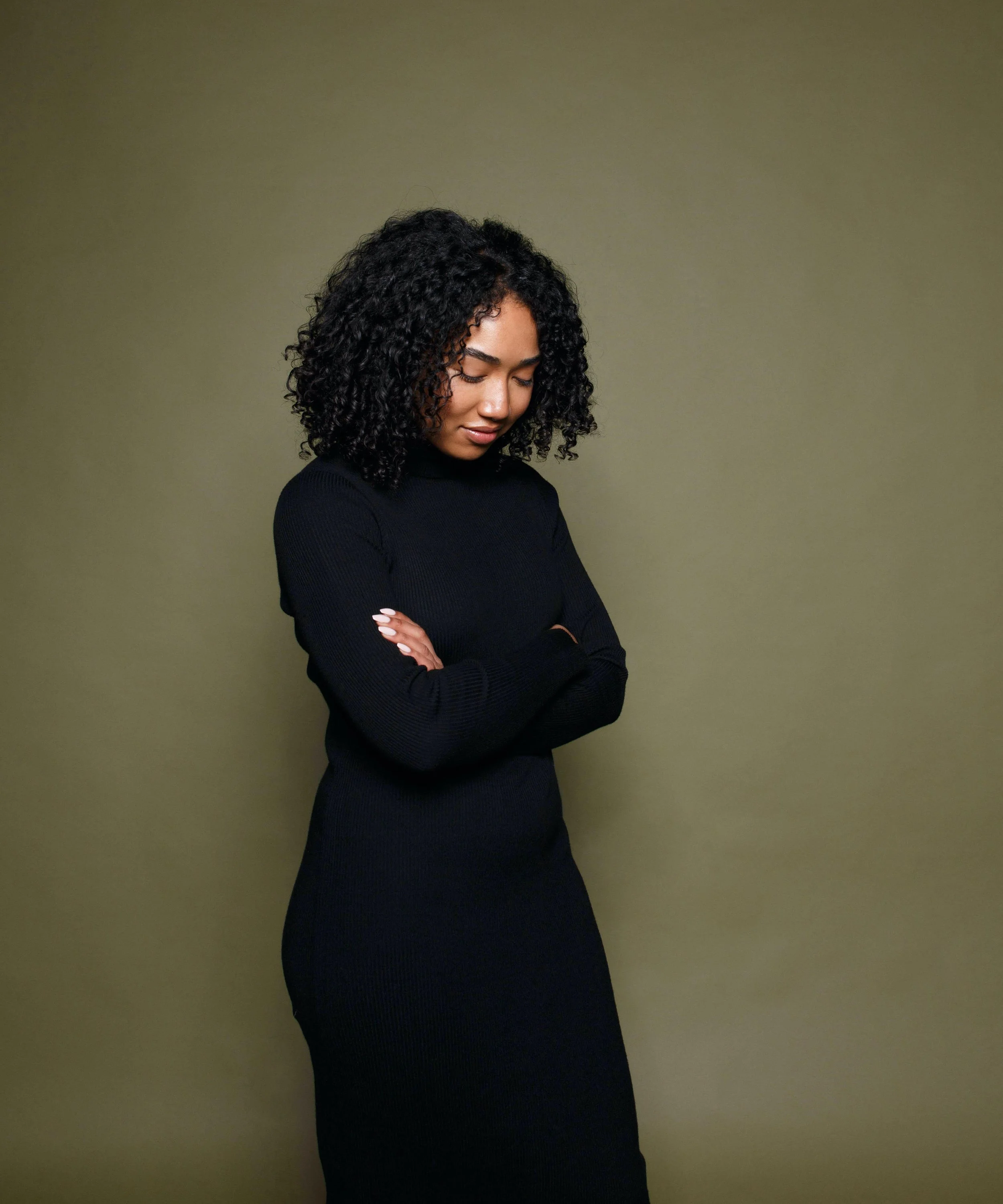 A woman with curly hair wearing a black dress with her arms crossed, standing against an olive-green background.