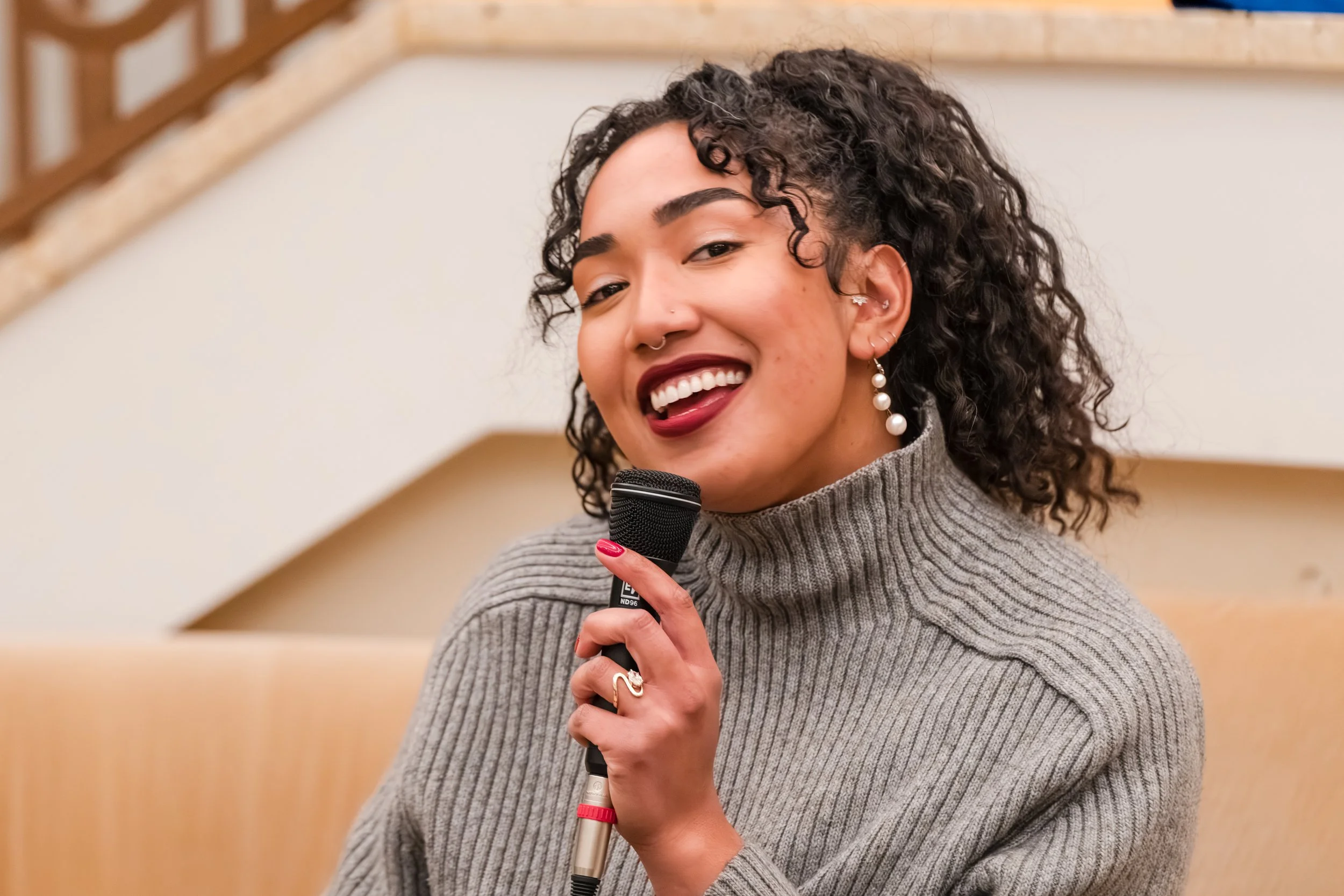 A young woman with curly hair, wearing a gray turtleneck sweater and pearl earrings, smiling while holding a microphone in her right hand.