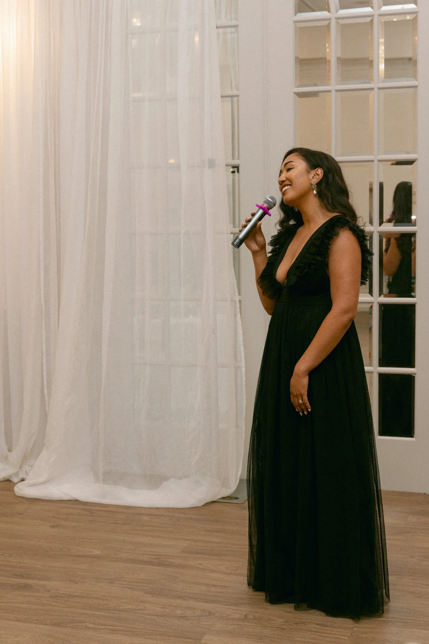 A woman in a long black dress holding a microphone, standing indoors near a white curtain and glass-paneled door, smiling and appearing to sing or speak.