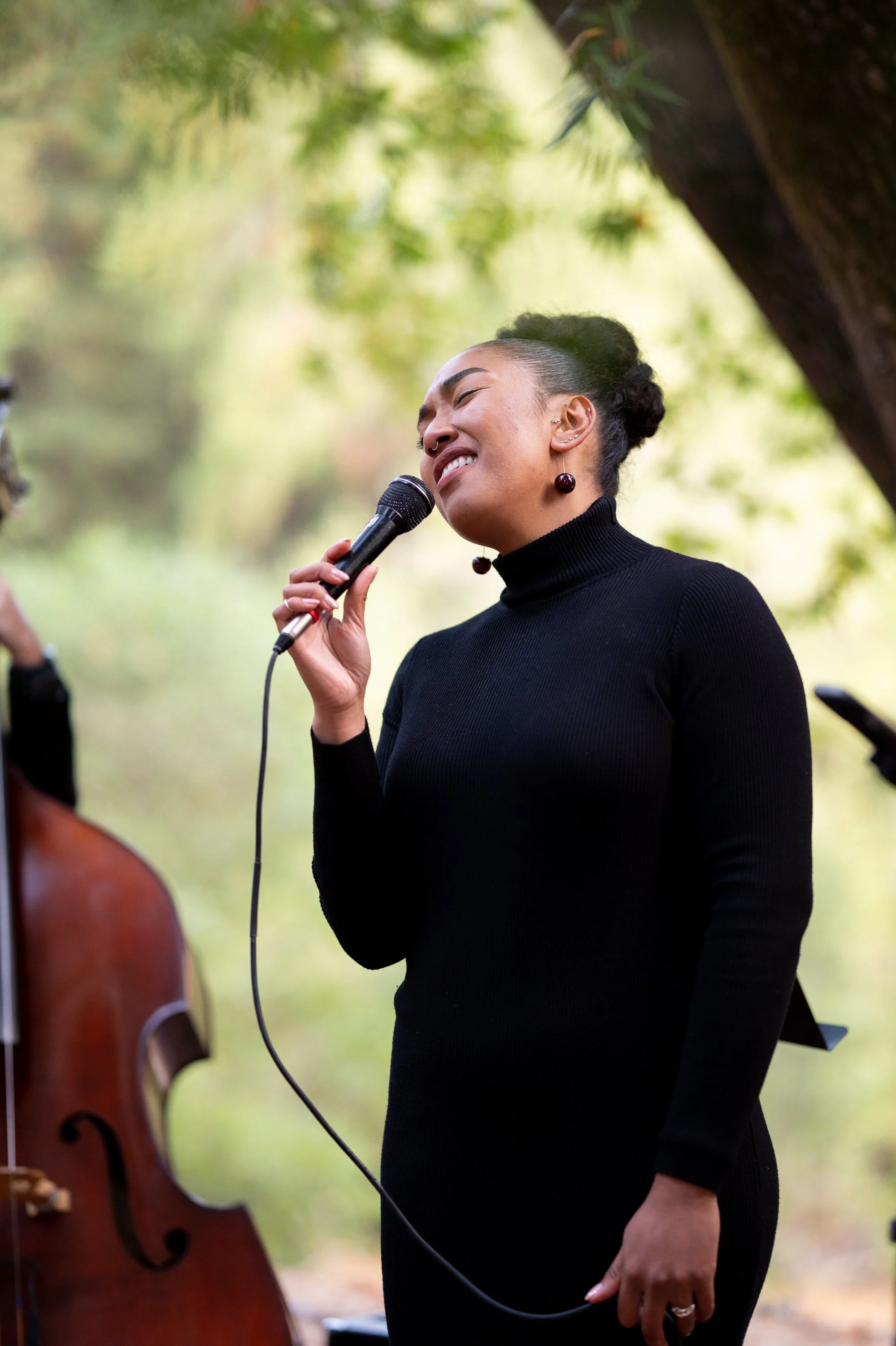 A woman in a black turtleneck dress singing passionately into a microphone outdoors, with a double bass visible in the background.