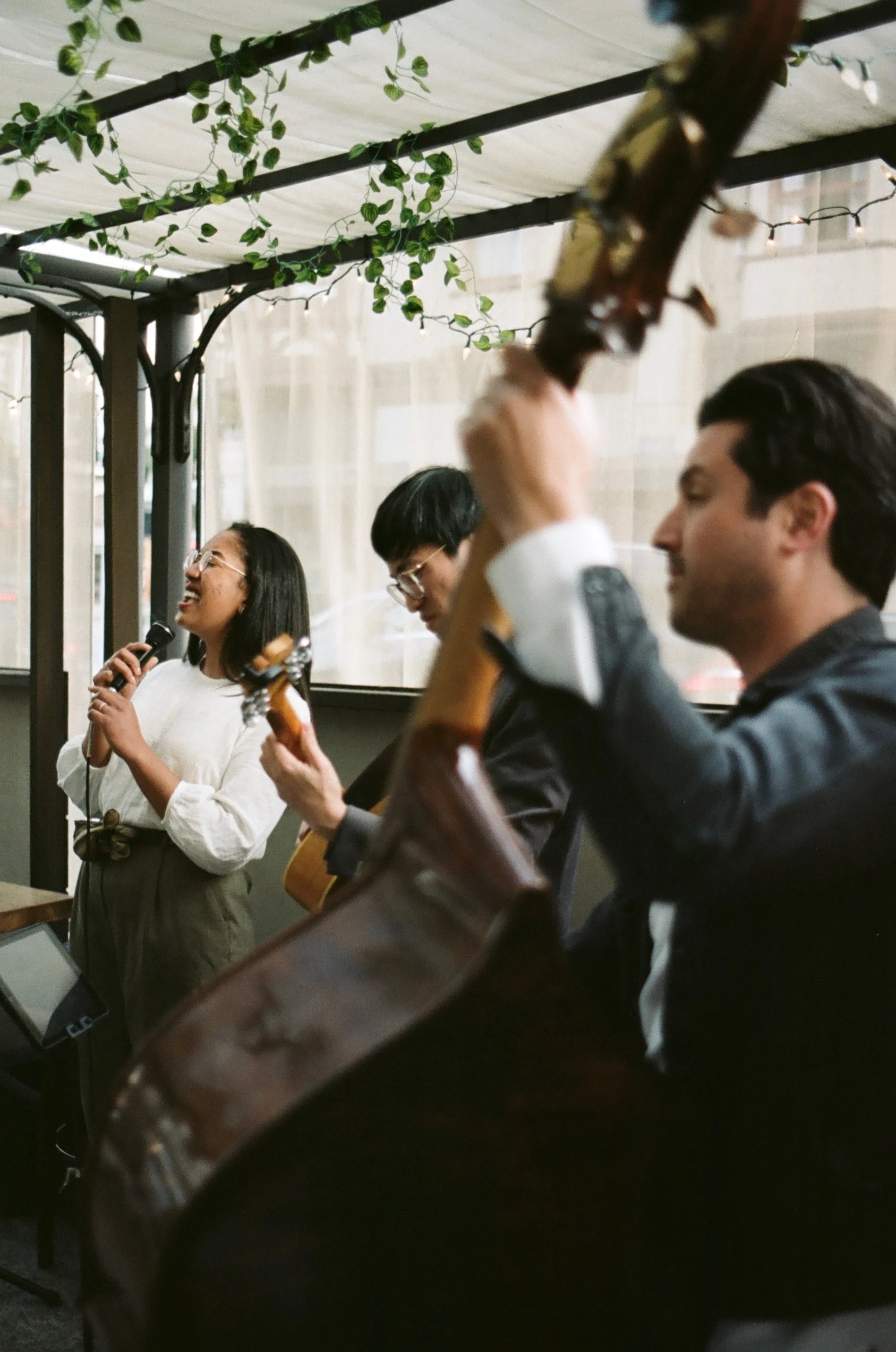 Group of three people performing music, with one woman singing into a microphone and two men playing guitars, in a cozy, decorated indoor setting.