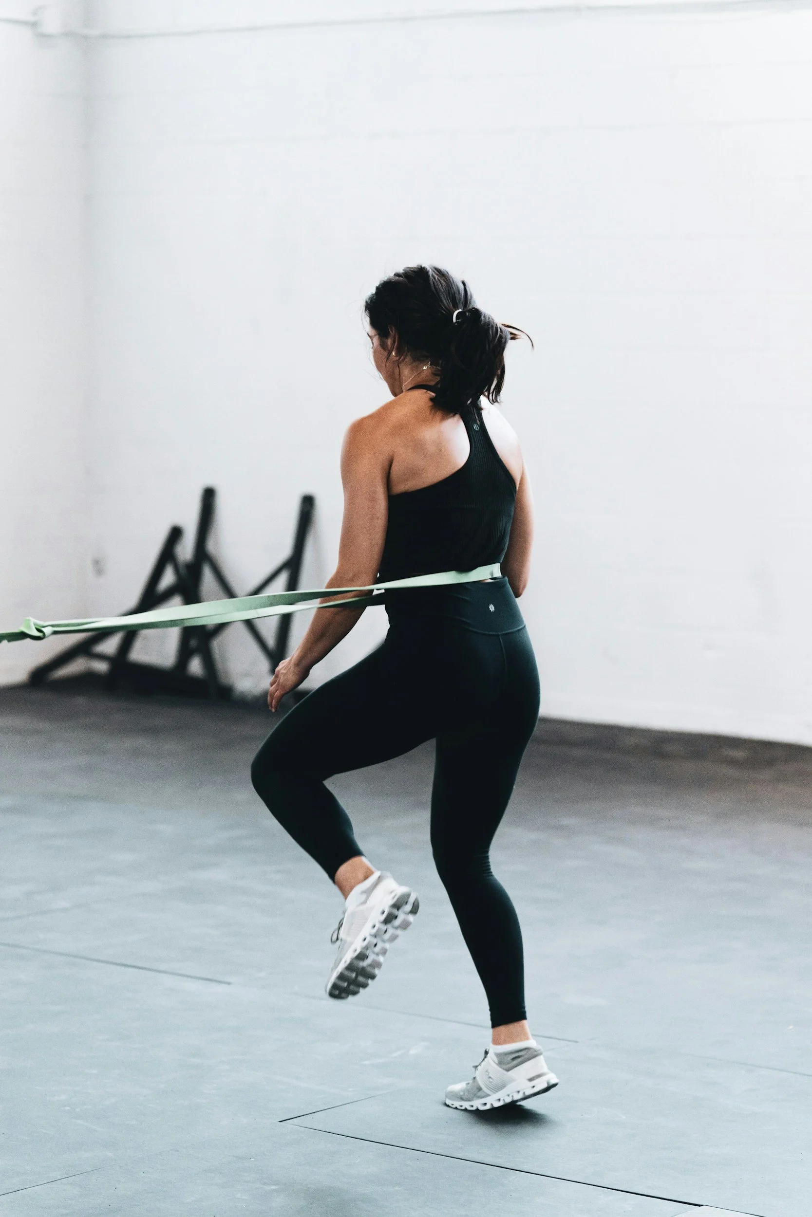 A woman in black athletic apparel and white sneakers jumping in a gym with gray flooring. She is holding a resistance band and has her hair tied back. In the background, there are three black chairs against a white wall.