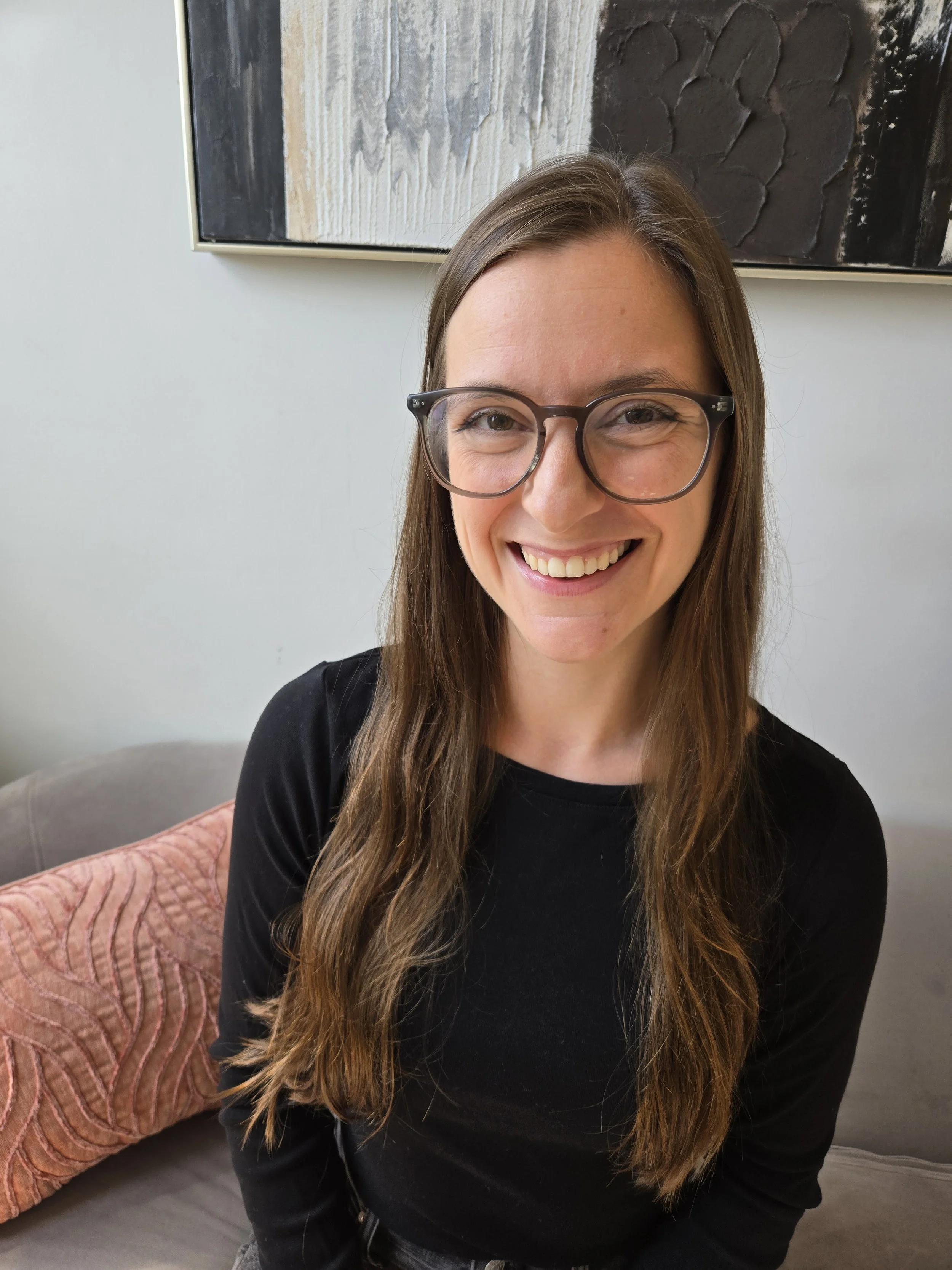 A woman with long brown hair, wearing glasses, a light gray turtleneck, and black pants, smiling indoors in front of green plants.