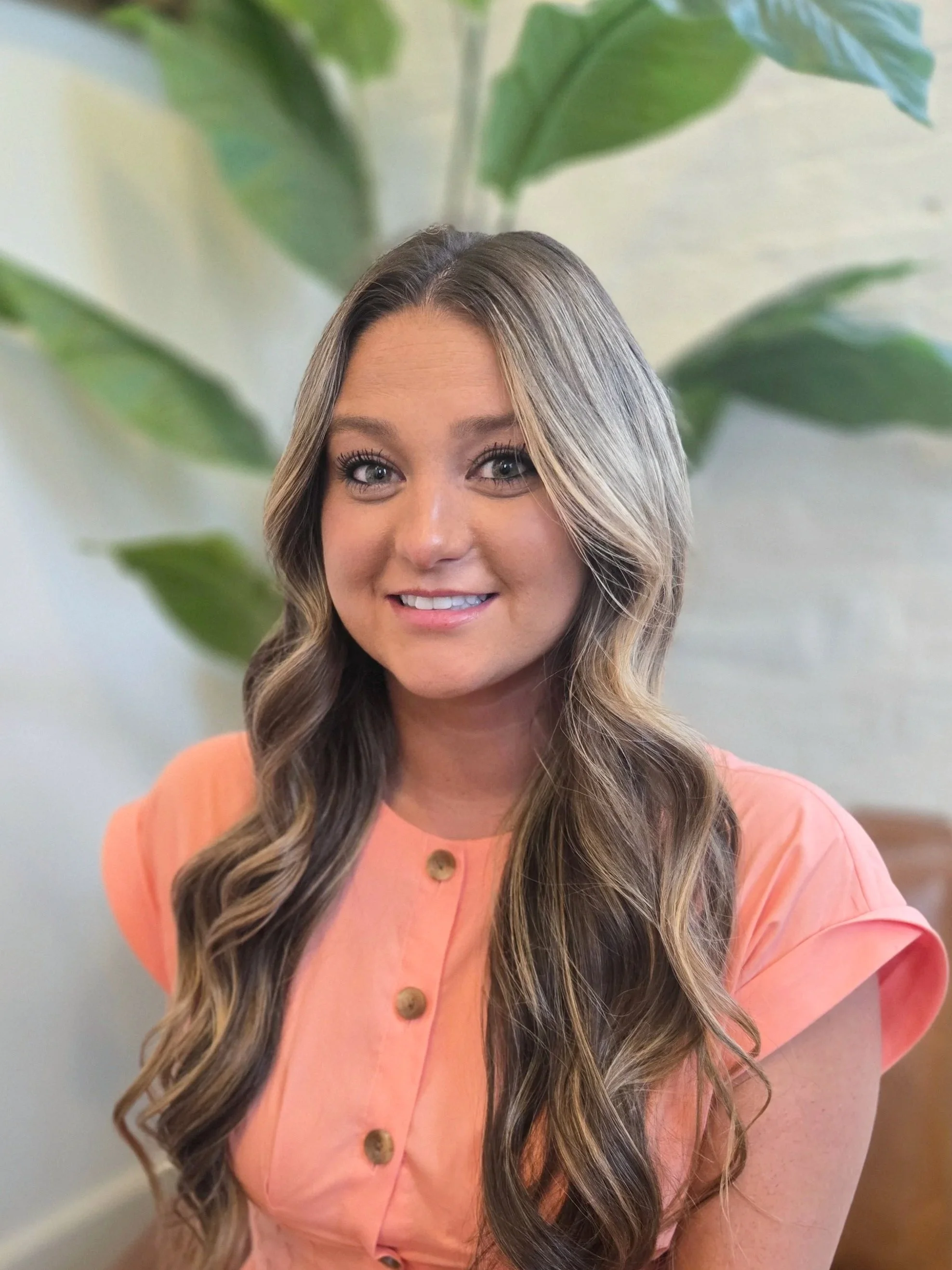 A woman with long, wavy brown hair and light makeup, smiling at the camera, wearing a white top and a gold necklace, with a potted succulent plant in the background.