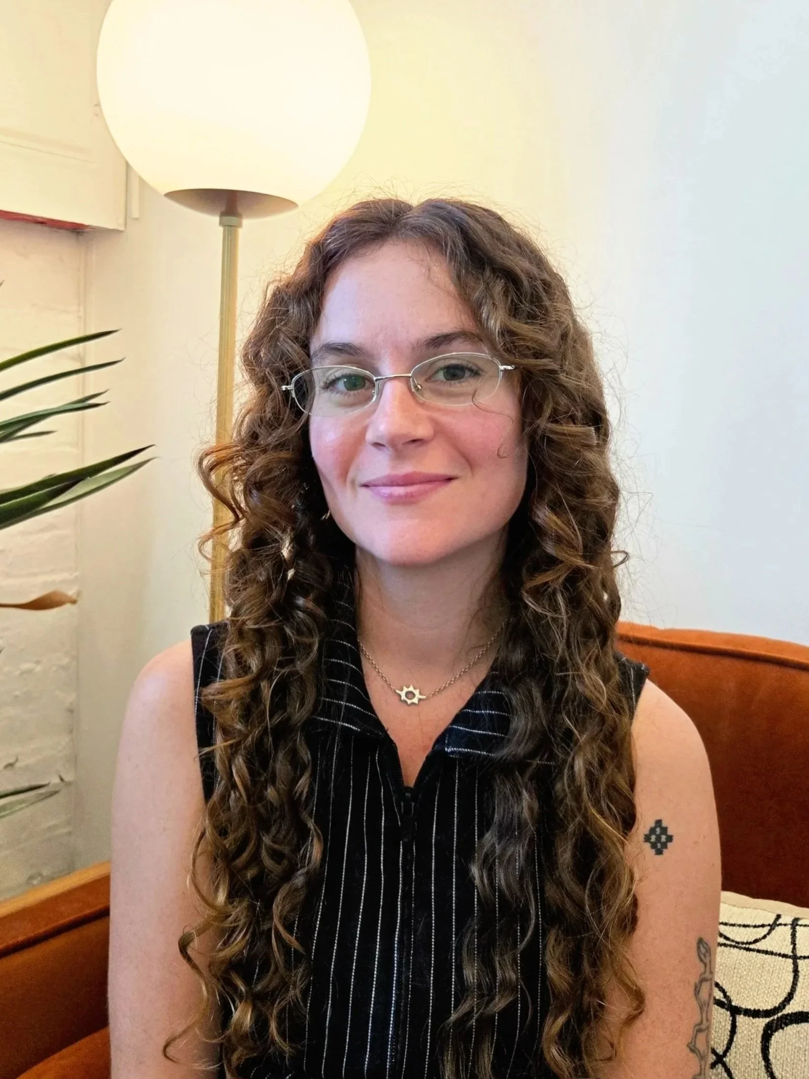 A woman with curly brown hair wearing glasses, a white button-up shirt, and a gold necklace with a gear pendant, sitting on a black couch against a white brick wall with a painting above.