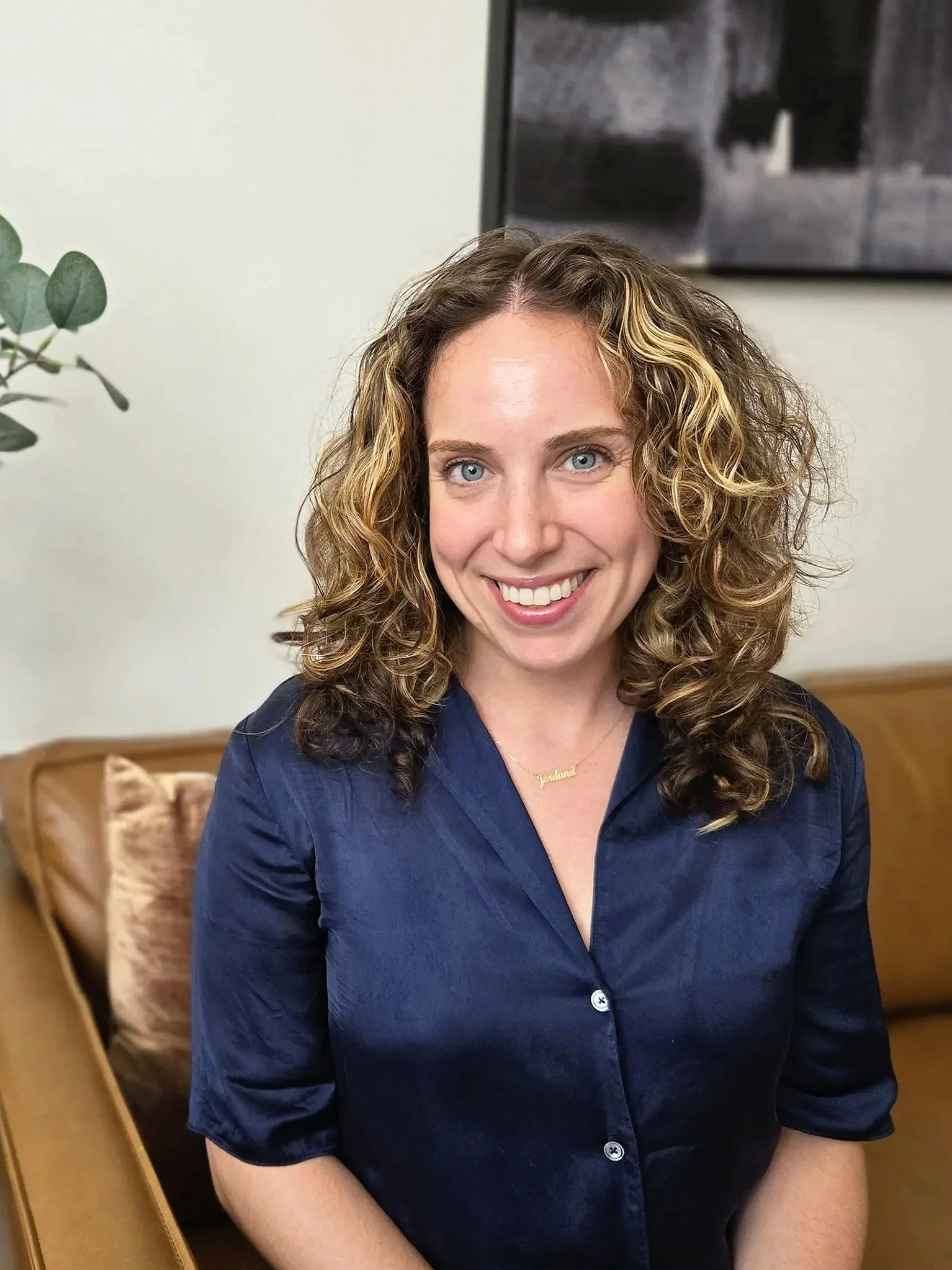 A smiling young woman with curly brown hair, wearing a black blazer, white shirt, and layered necklaces, sitting indoors.