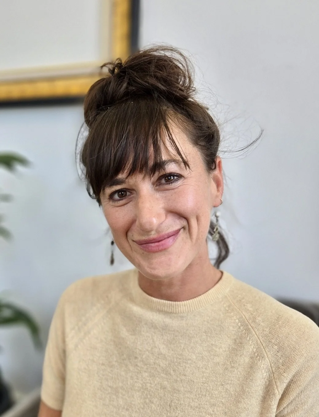 A woman with brown hair styled in a messy bun, wearing a beige sweater, smiling indoors with a white wall and a framed picture in the background.
