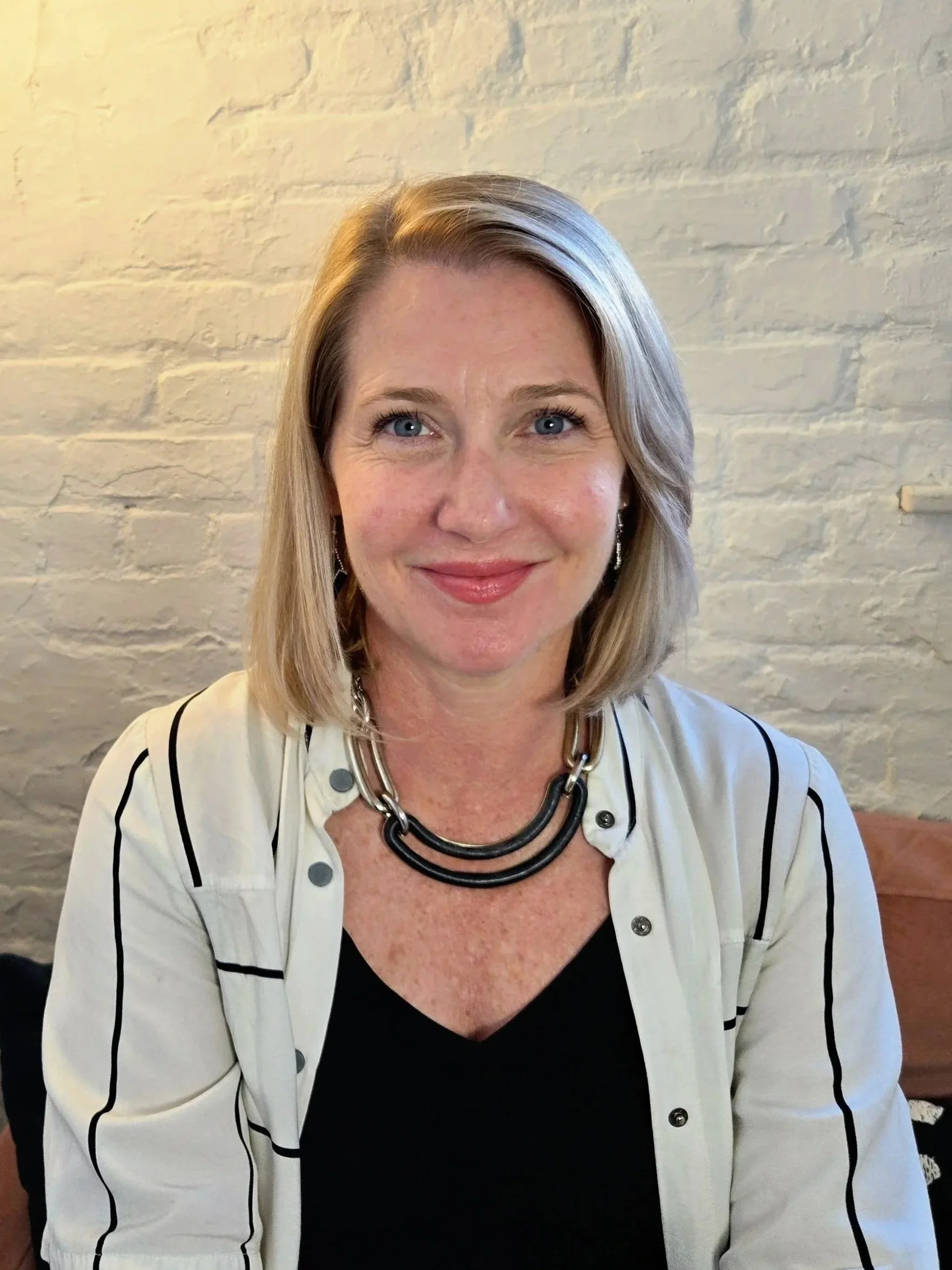 Close-up of smiling woman with blonde hair, wearing glasses, layered necklaces, and a black sleeveless top, indoors in natural light.