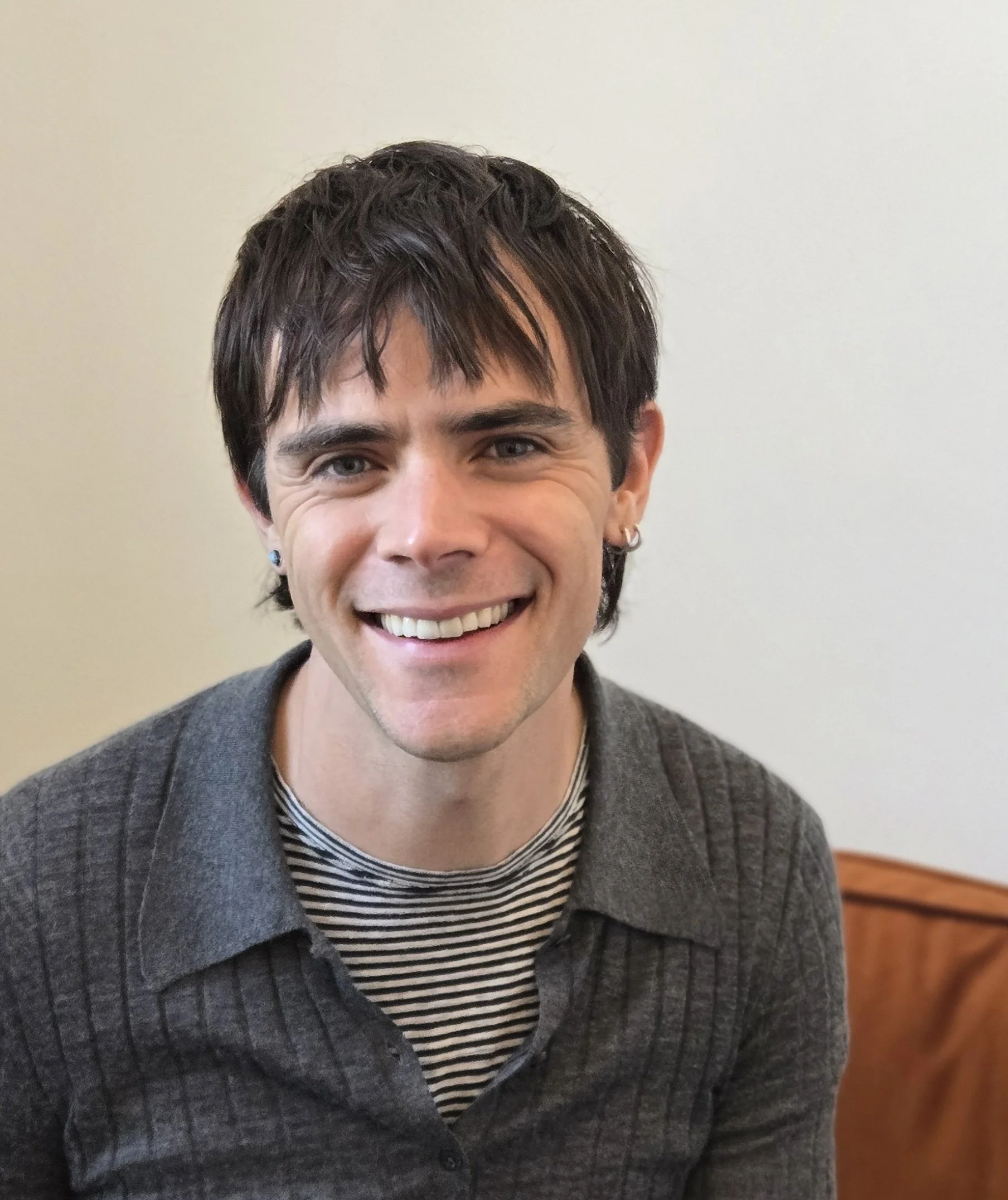 A young man with dark hair and earrings, smiling, wearing a gray cardigan over a striped shirt, sitting in front of a plain light-colored wall.