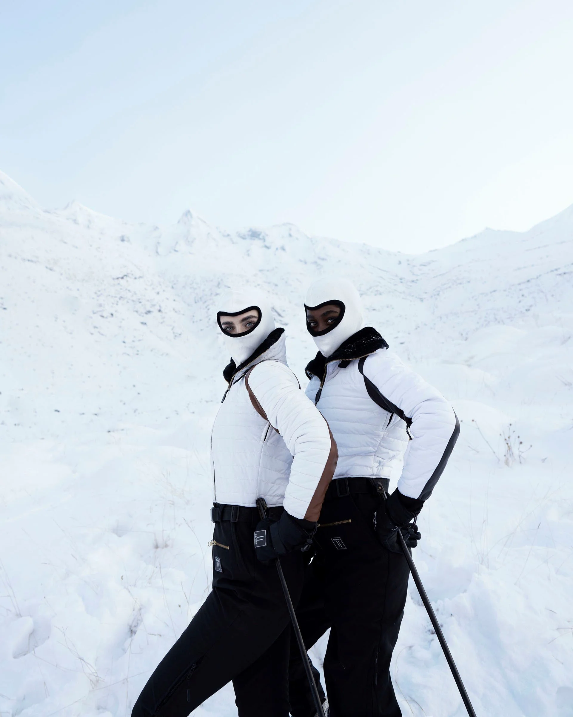 Two people in white winter jackets, black pants, and balaclavas stand in a snowy landscape, holding ski poles and looking at the camera.